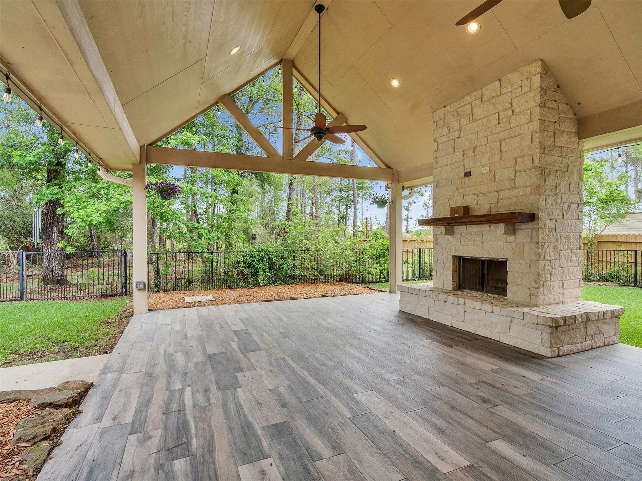 18005 Cindy Lane Conroe, TX 77302 - Photo 40 of 49 This photo showcases a spacious, covered outdoor patio with a high vaulted ceiling, modern wood-like tile flooring, and a large stone fireplace. It's surrounded by a lush, fenced backyard, perfect for relaxation and entertaining.