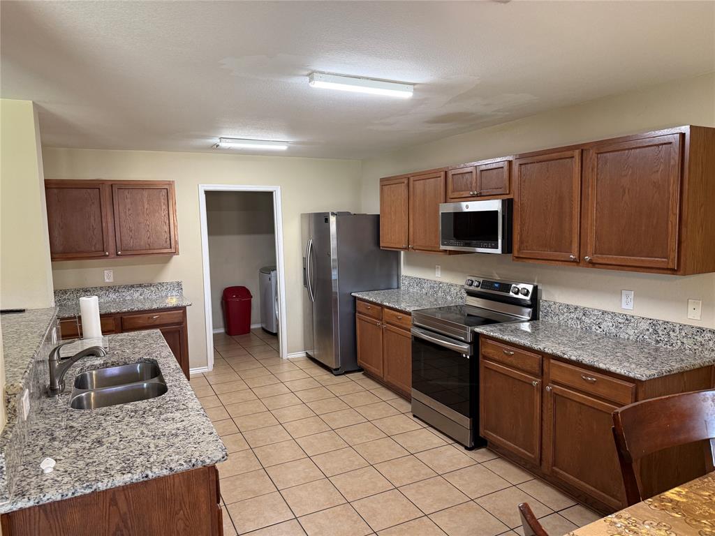 5718 Green Ivy Road Denton, TX 76210 - Photo 5 of 23 Kitchen featuring appliances with stainless steel finishes, light stone counters, brown cabinetry, light tile patterned floors, and a textured ceiling