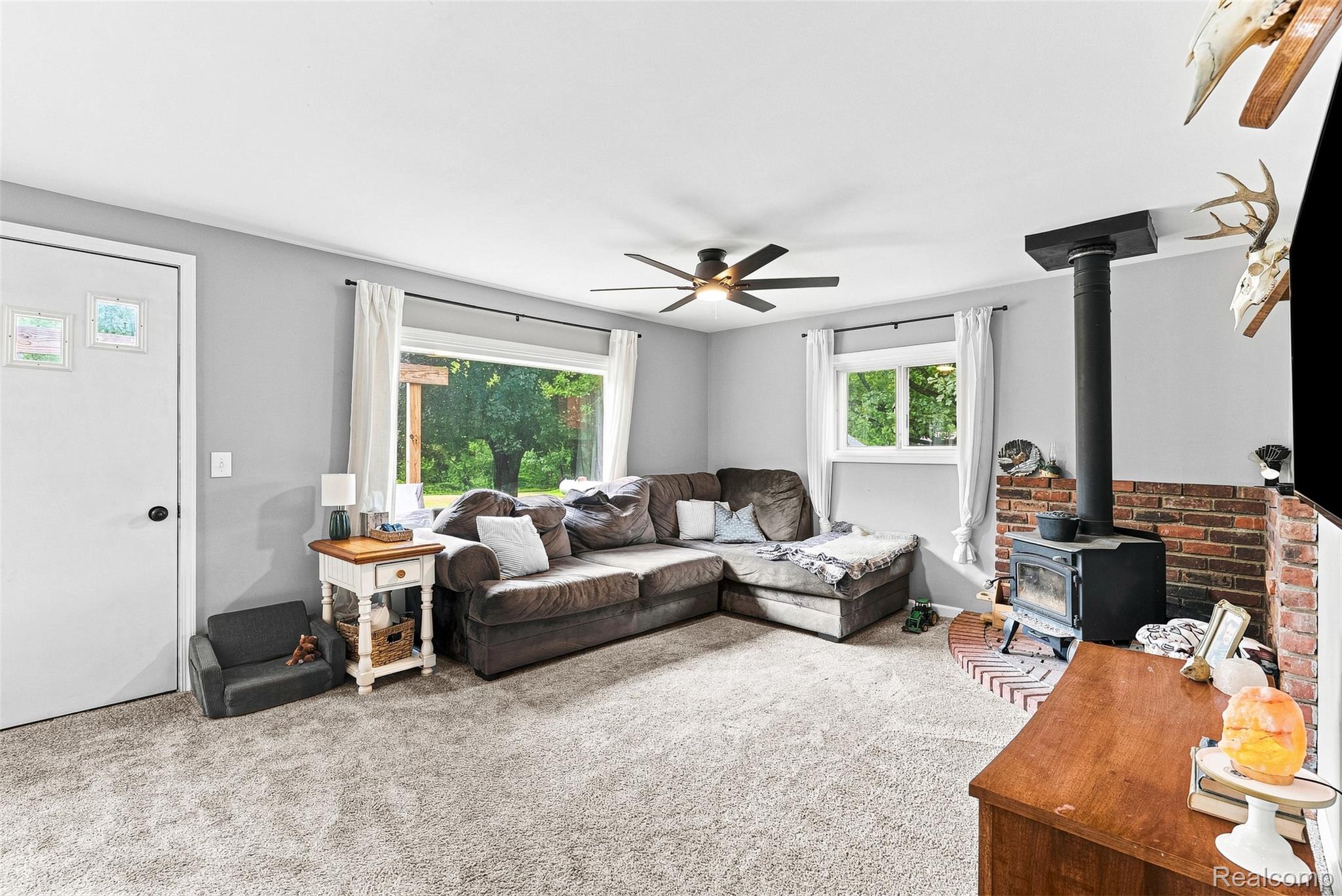 3319 Valentine Road Lapeer, MI 48446 - Photo 6 of 39 Spacious living area featuring light gray walls, plush carpeting, a ceiling fan with integrated lighting, a wood-burning stove with a brick surround, and large windows