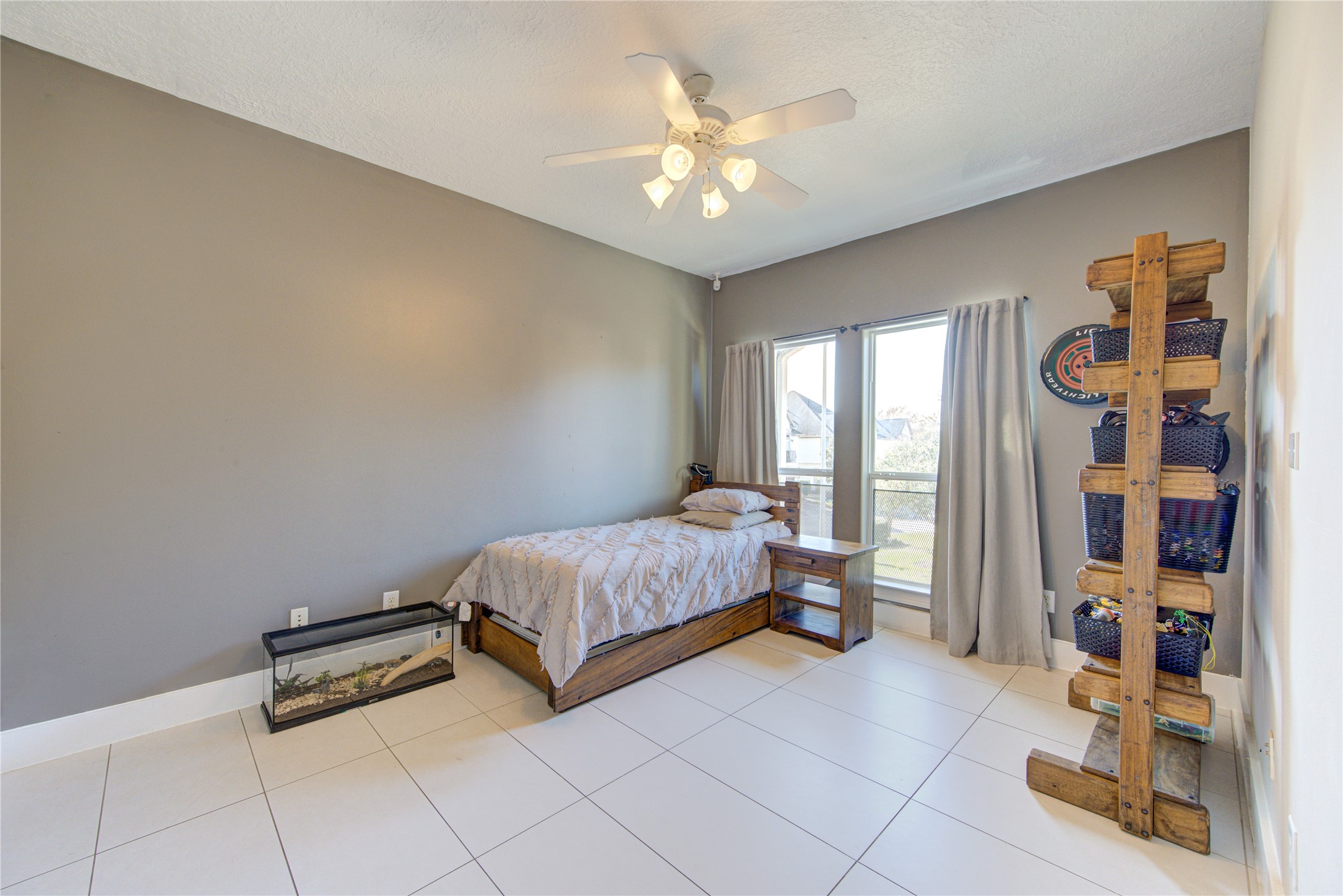 1419 Baldwin Square Lane Houston, TX 77077 - Photo 32 of 45 This room features a neutral color palette with a cozy single bed, large windows for natural light, and a ceiling fan. It has a tiled floor and ample storage space with a wooden shelf unit, creating a clean and functional environment.