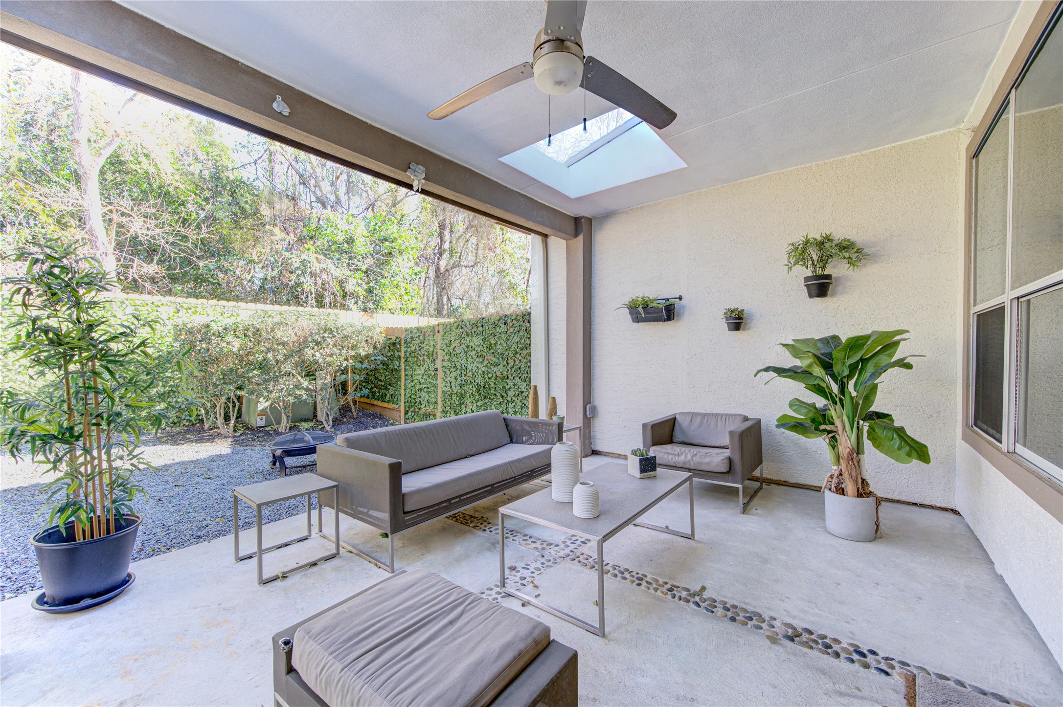 1419 Baldwin Square Lane Houston, TX 77077 - Photo 39 of 45 This photo showcases a modern, covered patio with comfortable seating, a ceiling fan, and a skylight. The space is accented with potted plants and pebble detailing, creating a serene outdoor living area surrounded by greenery.