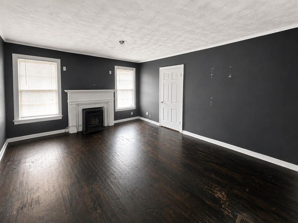 1010 Winters Street Dallas, TX 75216 - Photo 5 of 20 Spacious room featuring dark wood-finish flooring, a decorative fireplace with a white mantel, two windows with white trim, dark wall paint, and a white paneled door