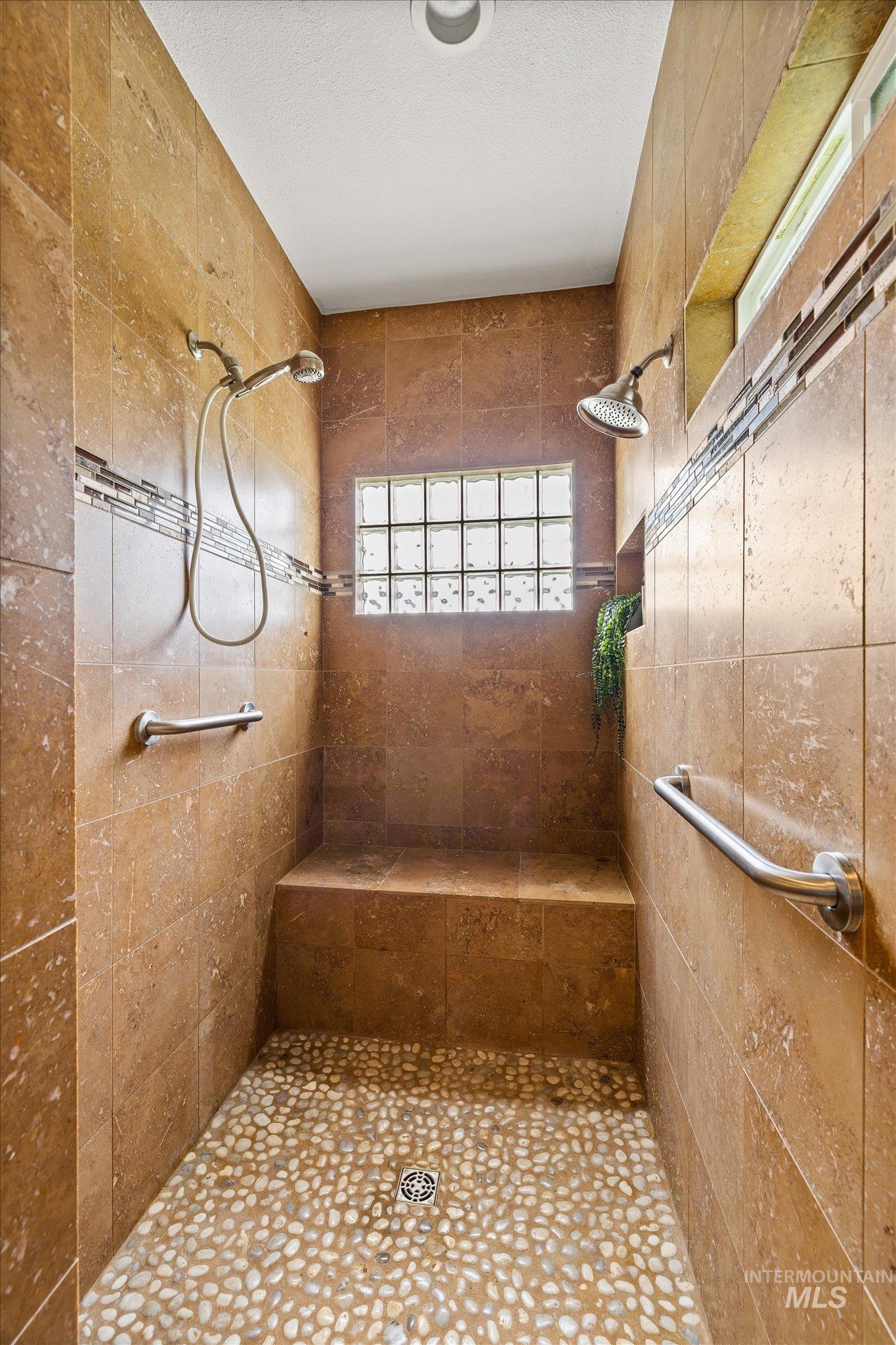 1140 Edgemont Road Emmett, ID 83617 - Photo 19 of 50 Spacious shower featuring dual showerheads, stone tile walls with decorative glass accents, a built-in bench, and a pebble-finish floor