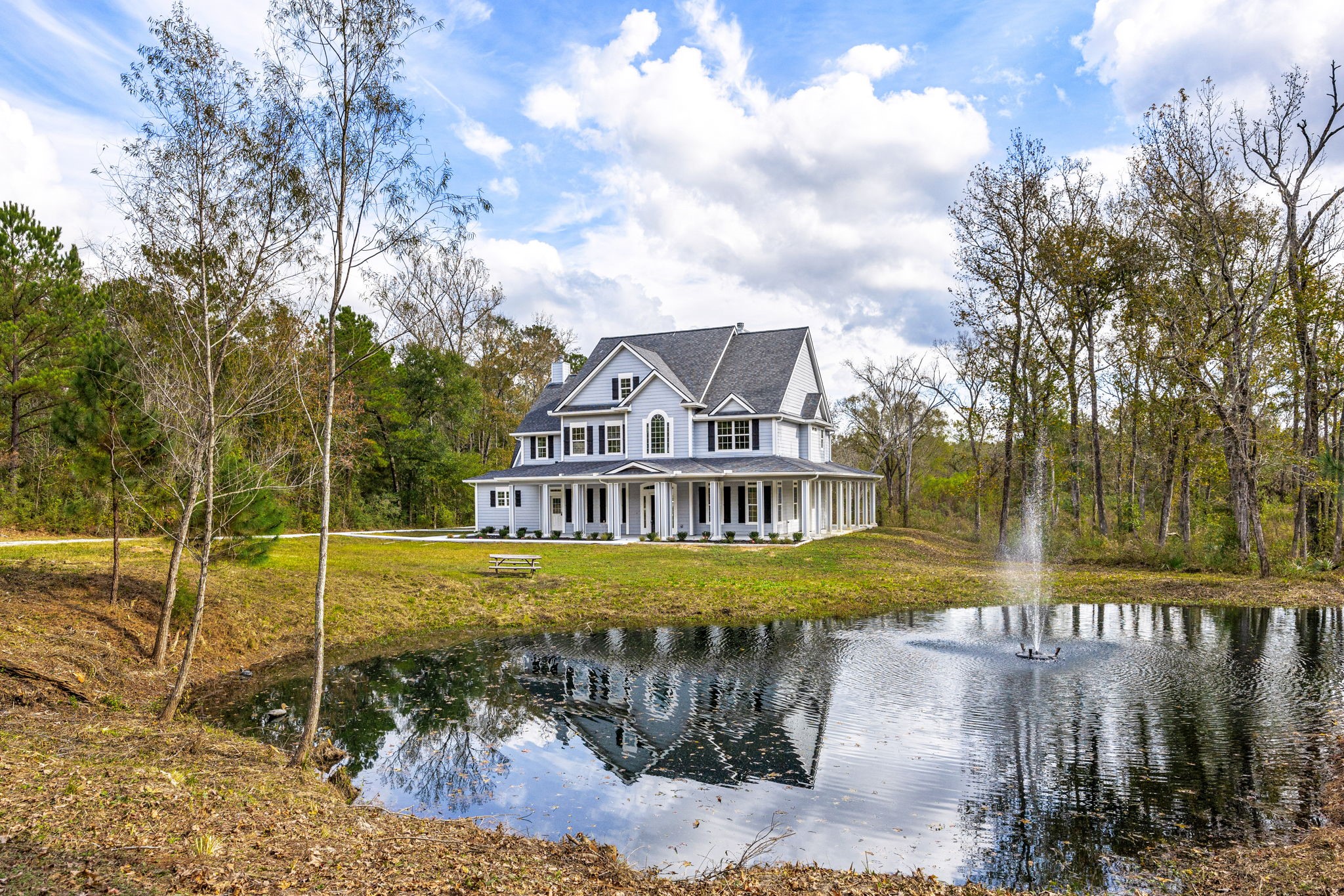 This photo showcases a charming, large two-story house with a wraparound porch, set in a tranquil wooded area. It features a picturesque pond with a fountain in the foreground, offering a serene, nature-filled setting.