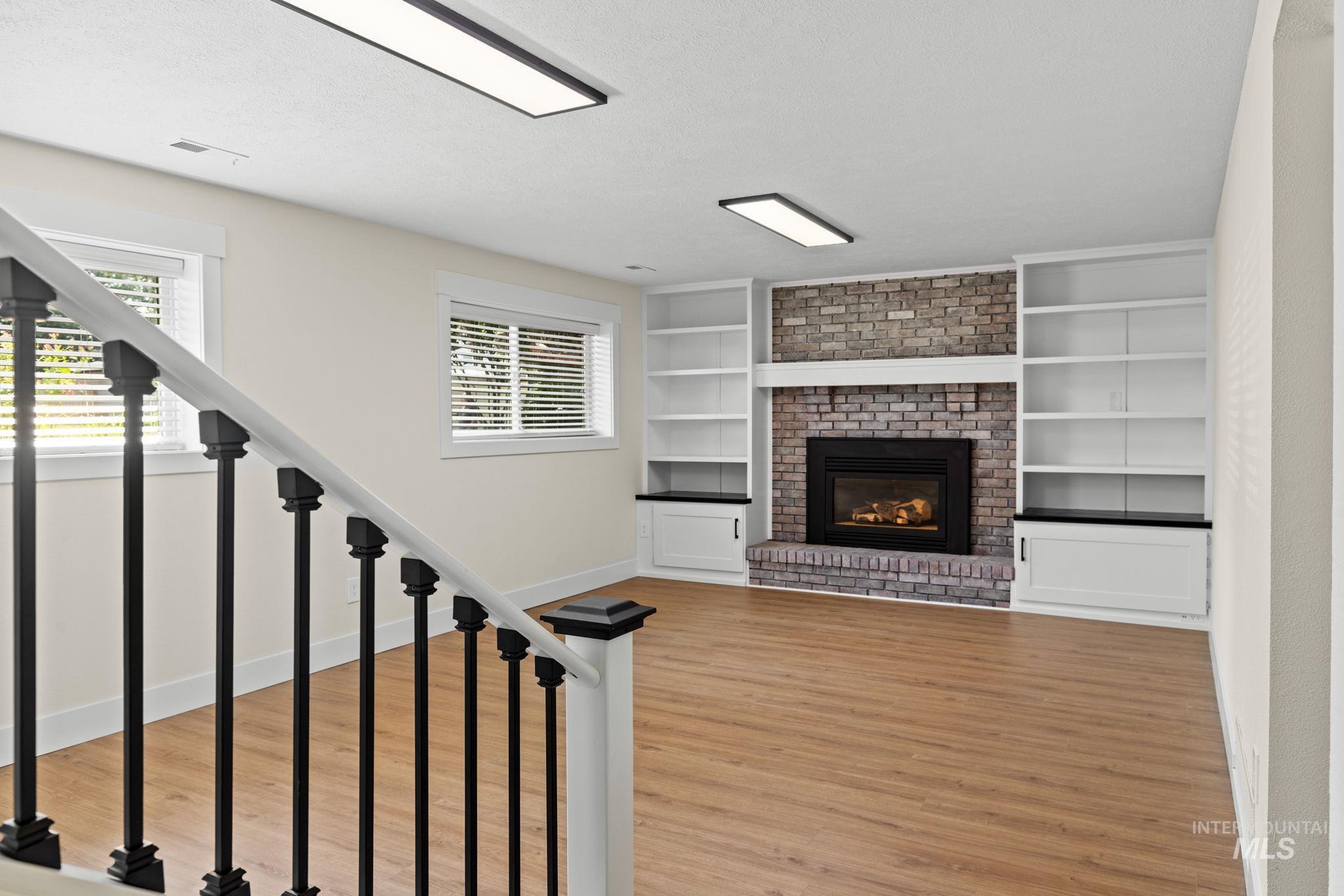 1675 Country Terrace Way Meridian, ID 83642 - Photo 25 of 50 Gas fireplace with brick surround, built-in shelving, white cabinetry with dark countertops, wood-finish flooring, and a staircase with black metal balusters