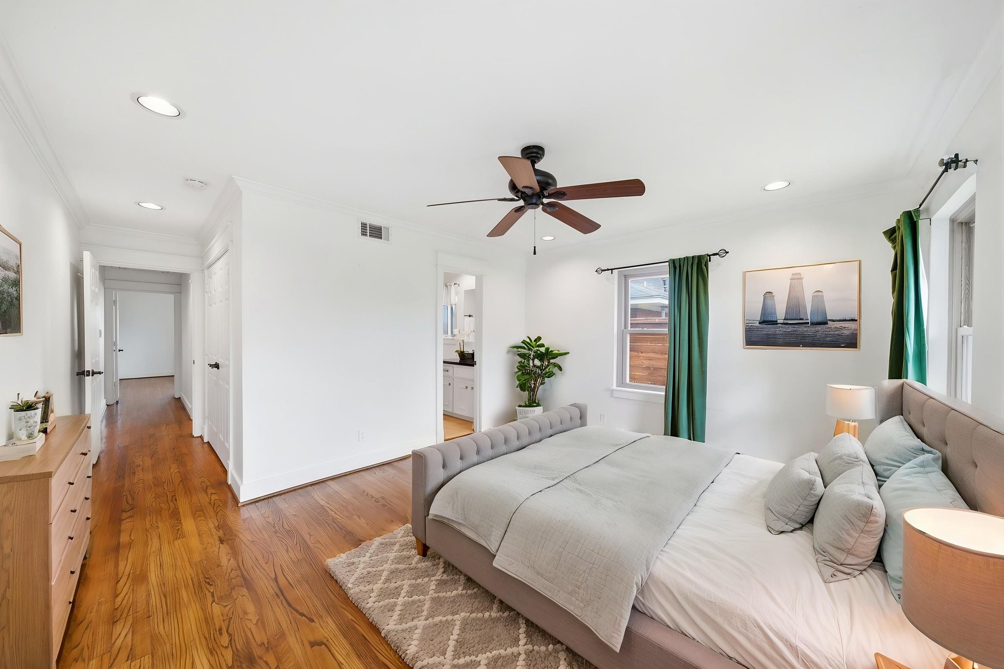 6427 Waltway Drive Houston, TX 77008 - Photo 21 of 37 *AI staged* This primary bedroom features a bright and airy atmosphere with hardwood floors. It includes a ceiling fan, recessed lighting, and ample natural light from three windows. There's also a glimpse into an adjoining bathroom and the common bedroom hallway.