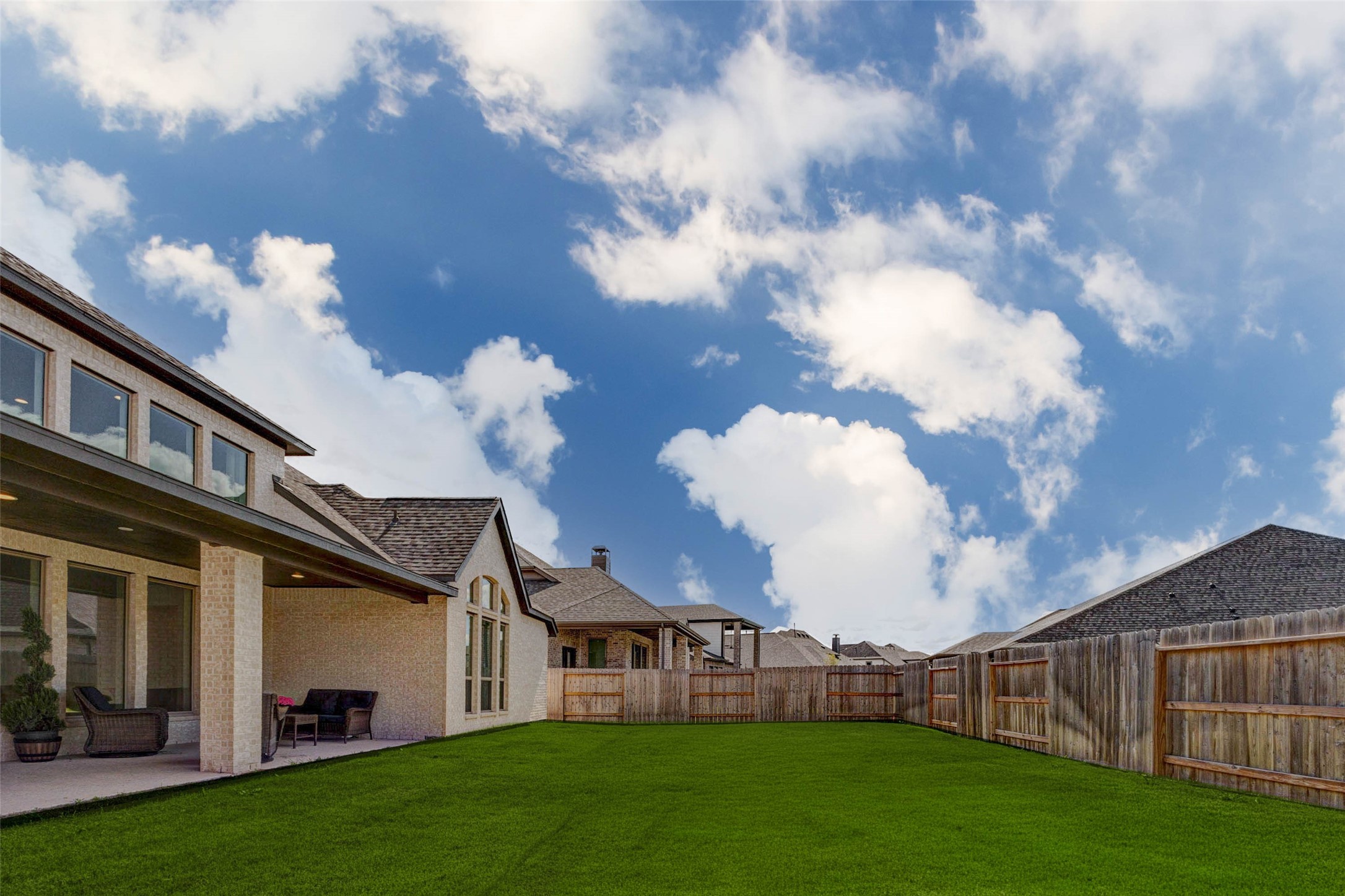 4322 Southpoint Way Fulshear, TX 77441 - Photo 44 of 50 Spacious backyard with a well-maintained lawn, bordered by a wooden fence. The home features a covered patio area, large windows, and a brick exterior. The sky is clear with scattered clouds, creating a bright and inviting atmosphere.