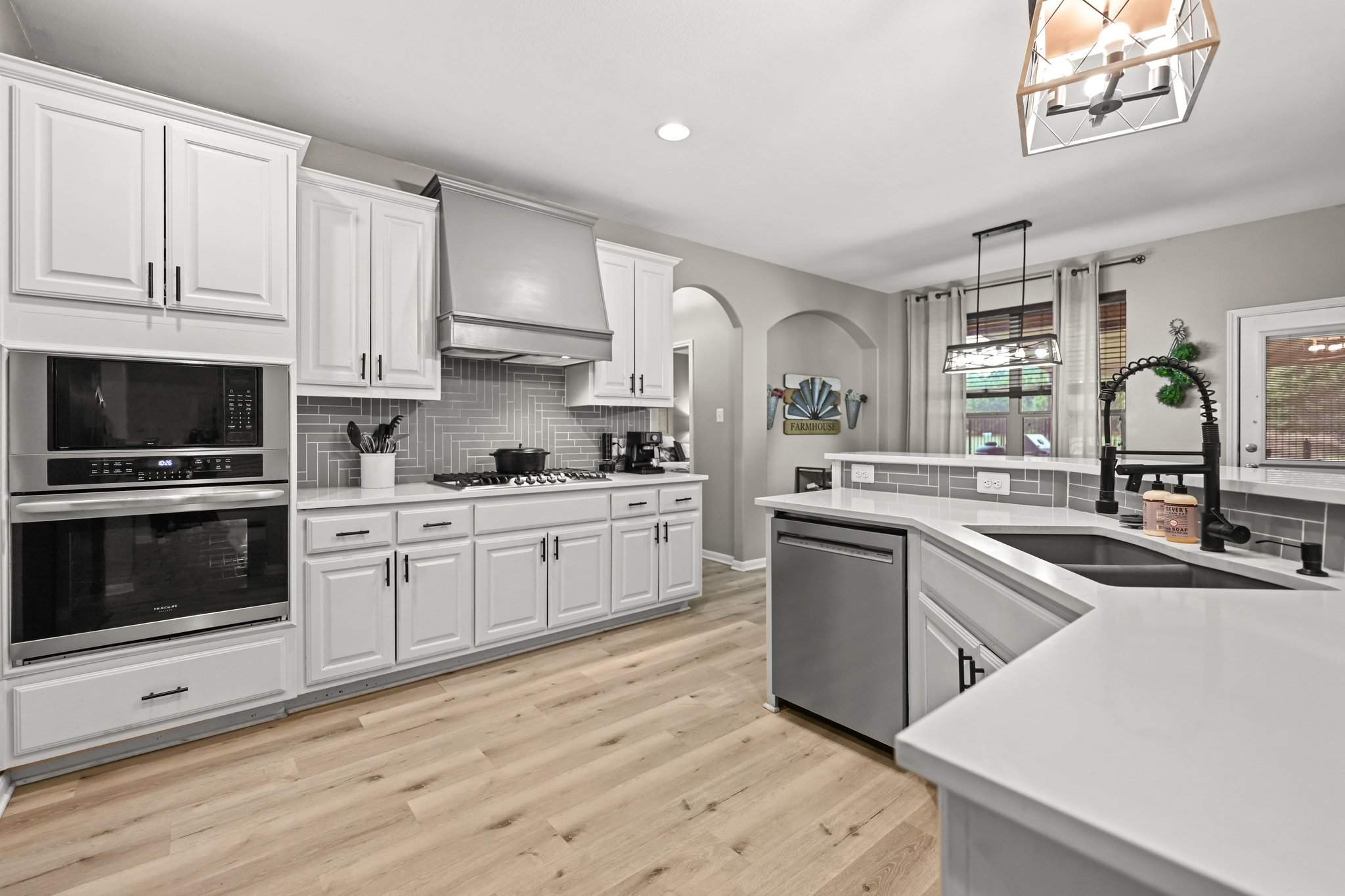 5528 Wild Foxglove Road Spicewood, TX 78669 - Photo 7 of 38 Modern kitchen featuring white cabinetry with black hardware, stainless steel appliances, light wood-finish flooring, a herringbone tile backsplash, and light-colored countertops