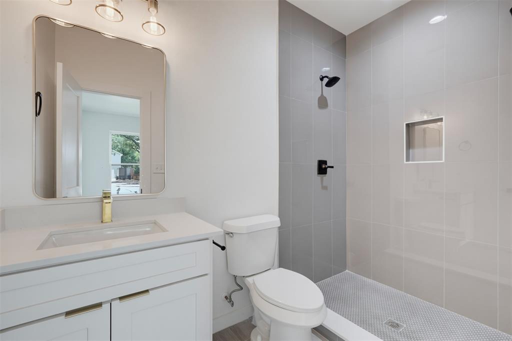 512 South O Connor Road Irving, TX 75060 - Photo 7 of 21 Modern bathroom featuring a walk-in shower with gray tile surround and black fixtures, a white vanity with an integrated sink and gold-tone faucet, and a contemporary wall-mounted mirror with overhead lighting