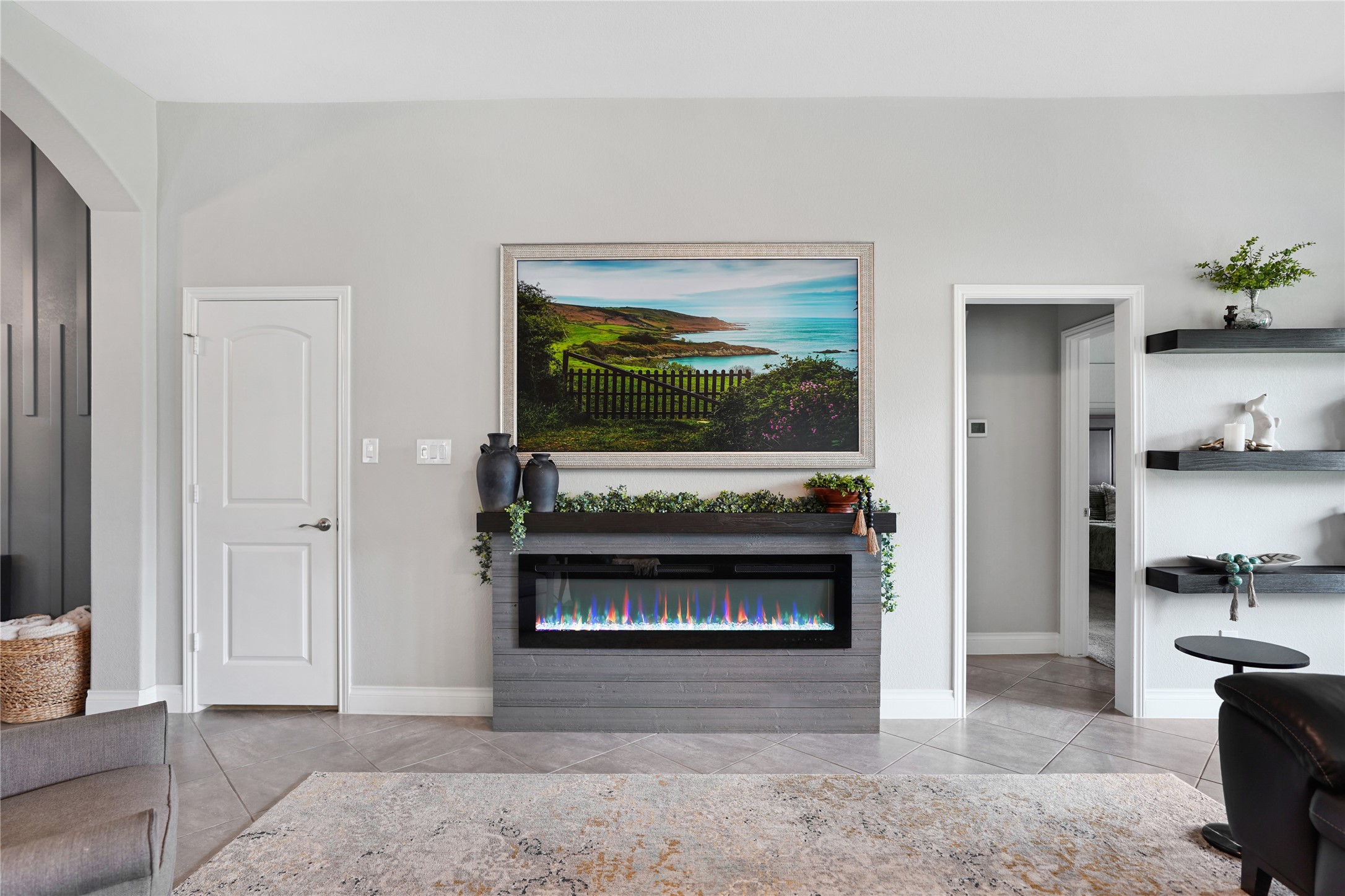 2345 Redwood Ridge Trail Manvel, TX 77578 - Photo 10 of 39 This modern living space features a sleek, wall-mounted electric fireplace beneath a vibrant landscape painting. The room has light gray walls, tiled flooring, and decorative shelving, creating a cozy and stylish atmosphere.