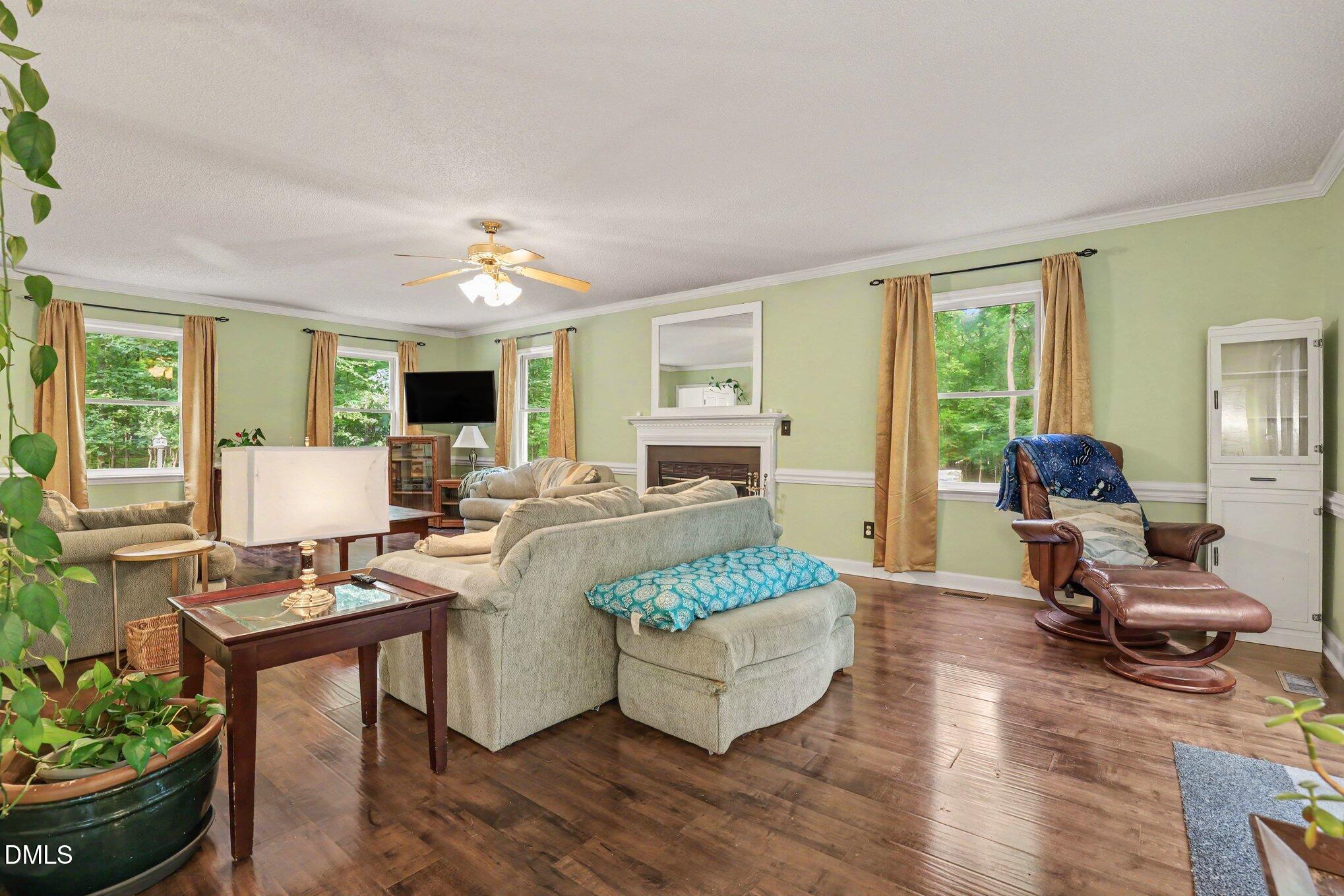 114 Oak Hollow Road Timberlake, NC 27583 - Photo 9 of 31 Spacious living area featuring wood-finish flooring, a fireplace with a white mantel, multiple windows with crown molding, and a ceiling fan