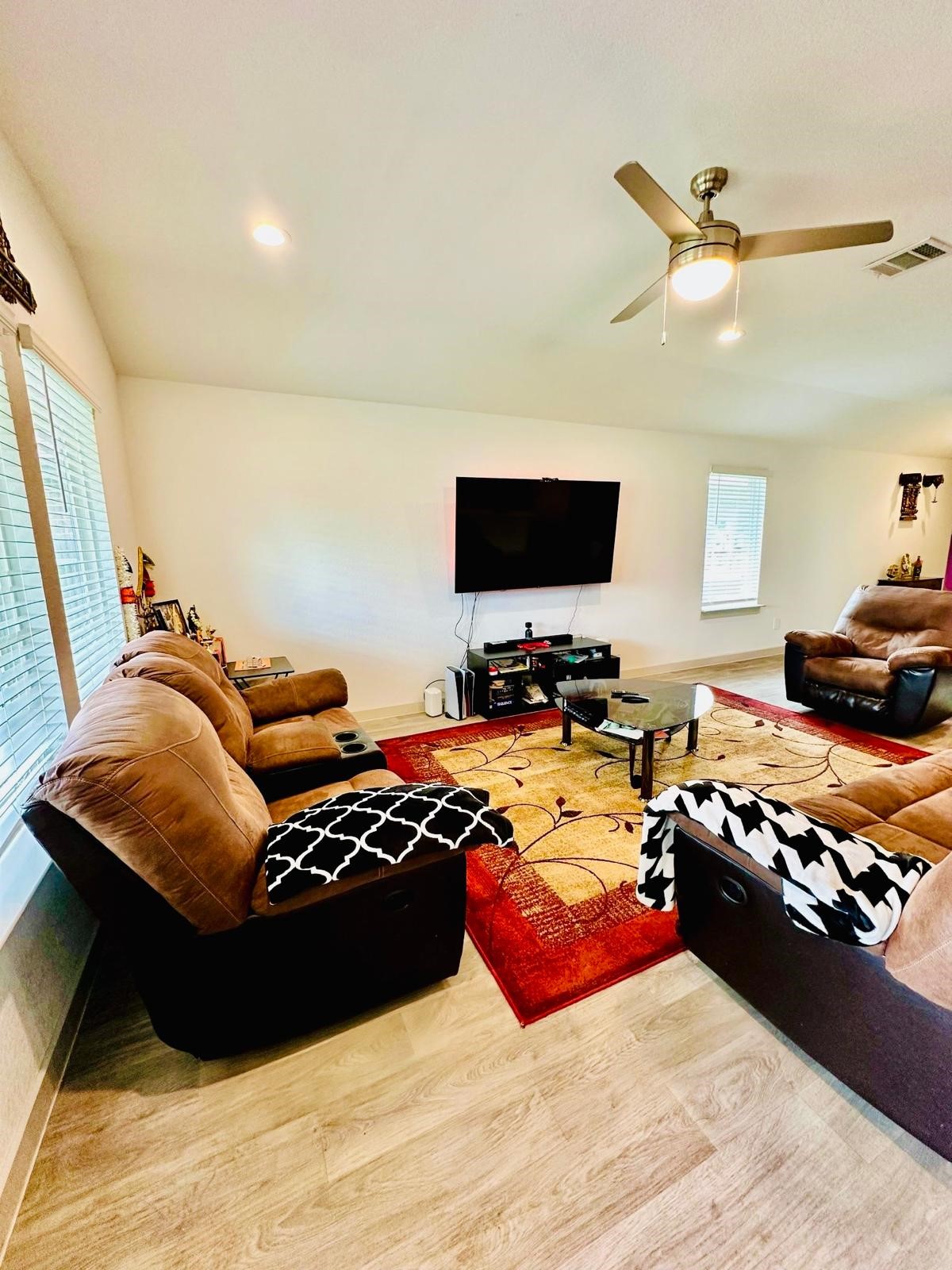 315 John House Lane Rosharon, TX 77583 - Photo 12 of 27 Spacious living room with cozy brown seating, a large wall-mounted TV, and stylish wood flooring. Natural light streams in through multiple windows, and a ceiling fan adds comfort.