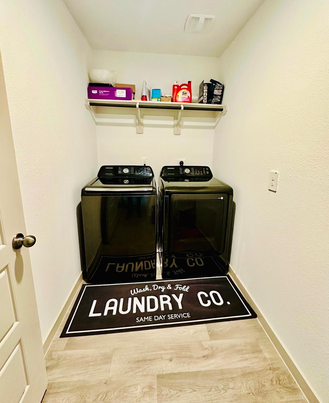 315 John House Lane Rosharon, TX 77583 - Photo 18 of 27 This photo shows a compact laundry room featuring modern black washer and dryer units. Above them, a shelf holds various laundry supplies. The room has clean white walls, light wood flooring, and a decorative mat with a laundry theme.