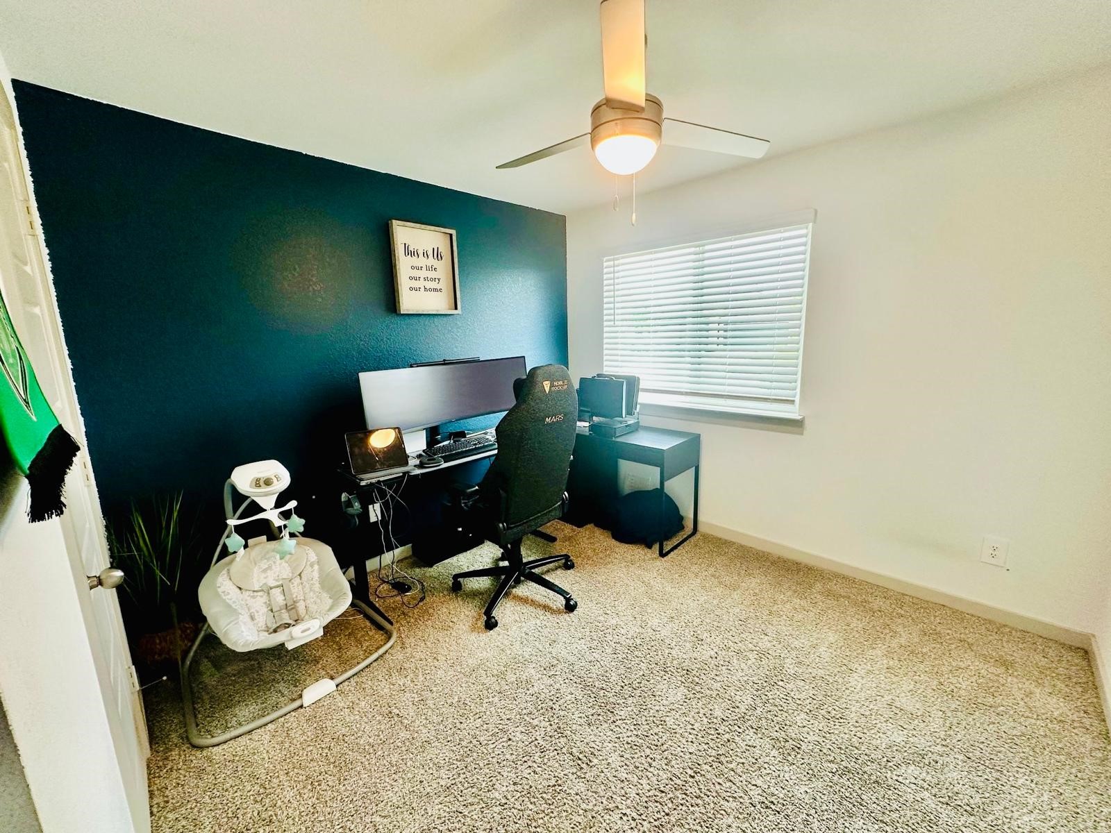 315 John House Lane Rosharon, TX 77583 - Photo 20 of 27 Cozy home office with a modern ceiling fan, plush carpet, and a feature wall painted dark blue. It includes a desk setup with dual monitors and a comfortable chair, plus a window providing natural light.