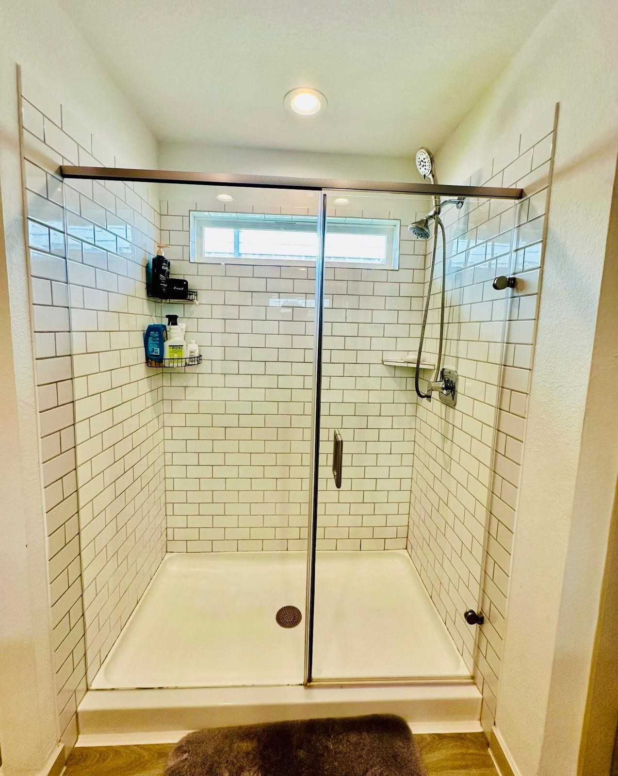 315 John House Lane Rosharon, TX 77583 - Photo 22 of 27 Modern walk-in shower featuring subway tile walls, a glass door, and a rain showerhead. It includes a small window for natural light and built-in shelves for toiletries.