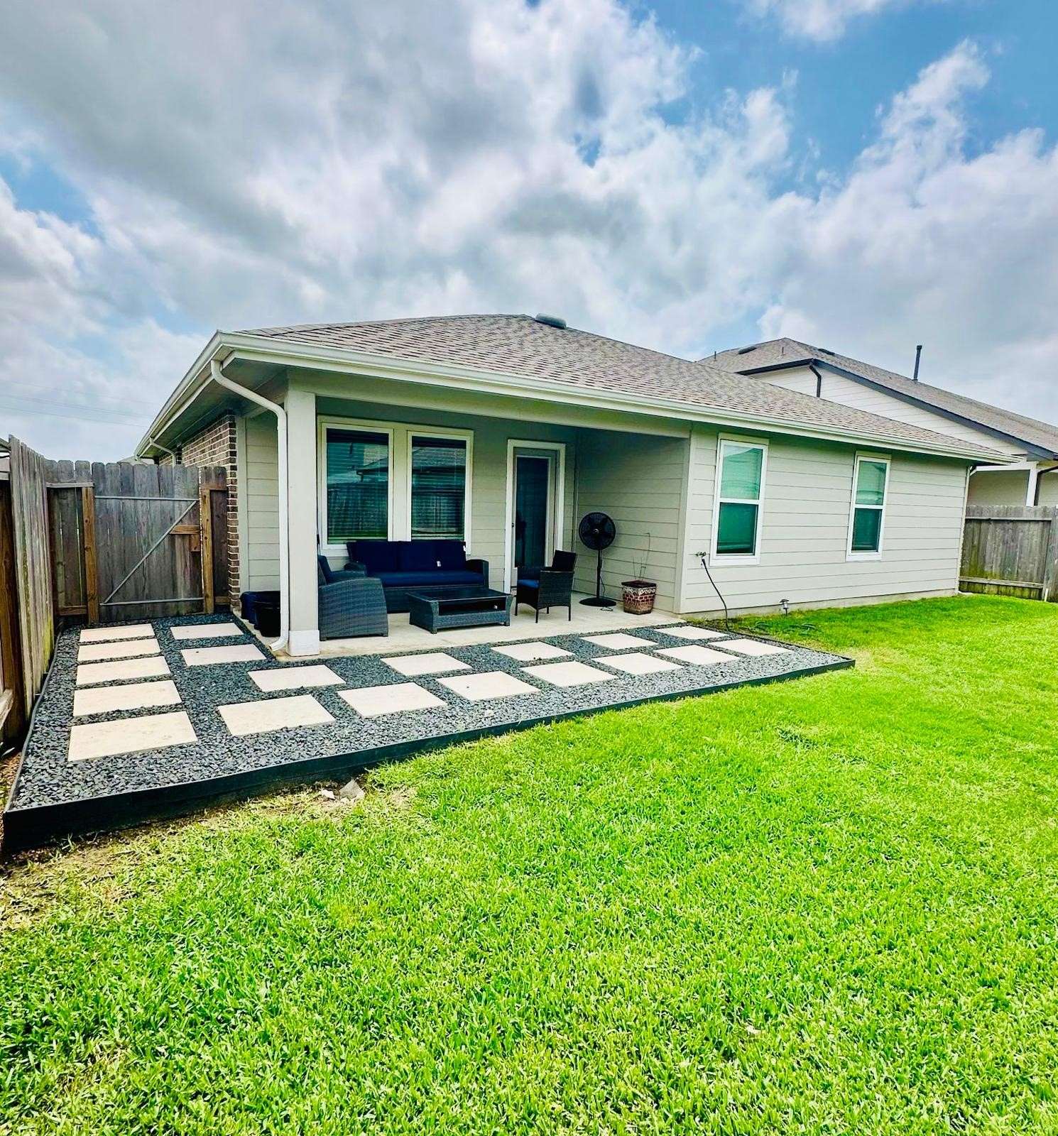 315 John House Lane Rosharon, TX 77583 - Photo 27 of 27 This photo shows the backyard of a home with a covered patio. It features outdoor seating on a checkerboard-style stone area, surrounded by grass, and enclosed by a wooden fence. The space is ideal for relaxing or entertaining.
