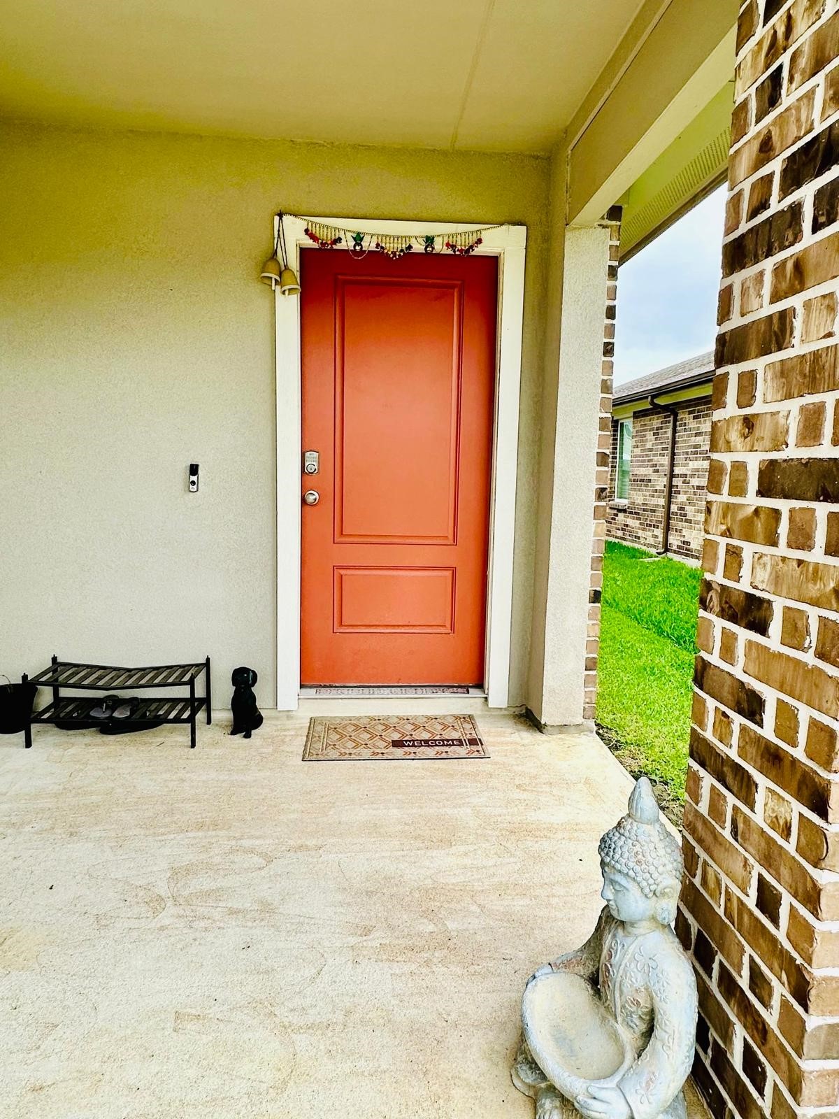 315 John House Lane Rosharon, TX 77583 - Photo 5 of 27 Front entryway featuring a bold red door, a welcoming mat, and decorative elements like a small bench and a serene statue. The area is framed by brickwork and opens to a lush green lawn, creating a warm, inviting atmosphere.