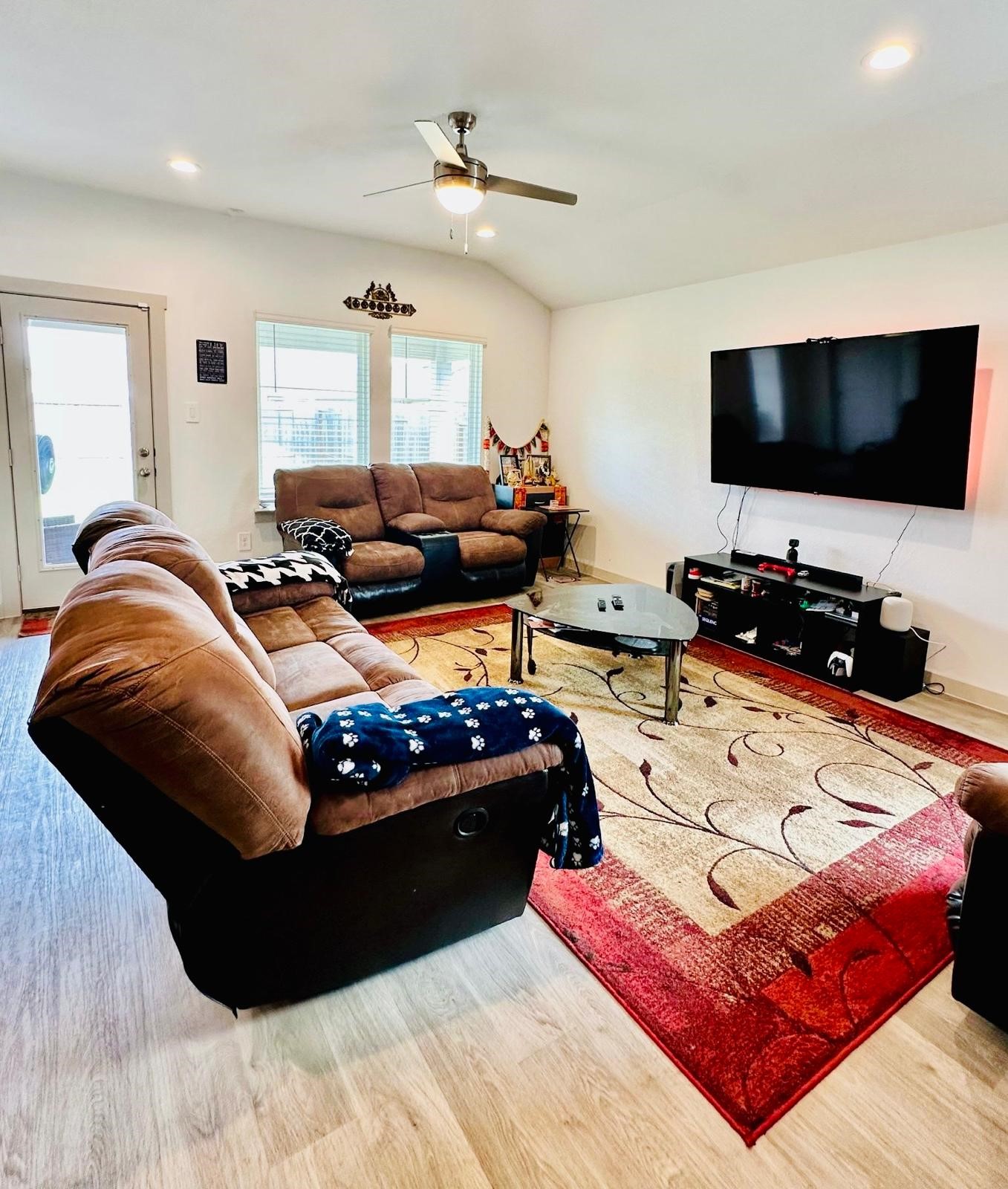 315 John House Lane Rosharon, TX 77583 - Photo 10 of 27 Spacious living room with cozy brown seating, a large wall-mounted TV, and stylish wood flooring. Natural light streams in through multiple windows, and a ceiling fan adds comfort.