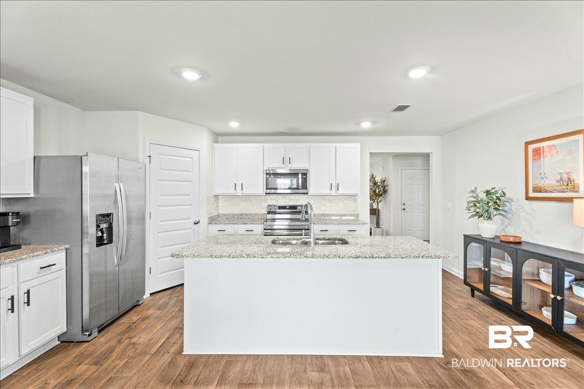 23641 Unbridled Loop Daphne, AL 36526 - Photo 12 of 40 Modern kitchen featuring an expansive center island with an undermount sink, white cabinetry, speckled countertops with a tile backsplash, stainless steel appliances, and wood-finish flooring