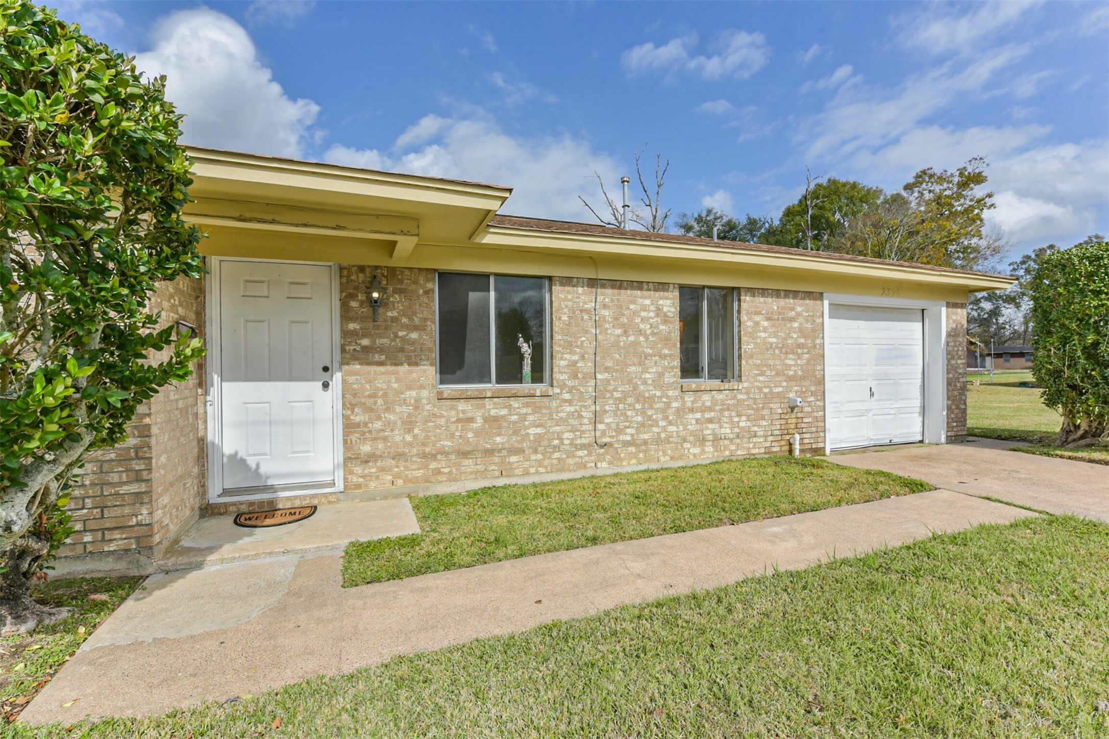 2390 Sarah Street Beaumont, TX 77705 - Photo 3 of 42 This photo shows a single-story brick home with a white front door and a single-car garage. It features a small front porch, a welcome mat, and a neatly maintained lawn with some shrubs. The home appears well-kept and is set under a blue sky with some clouds.