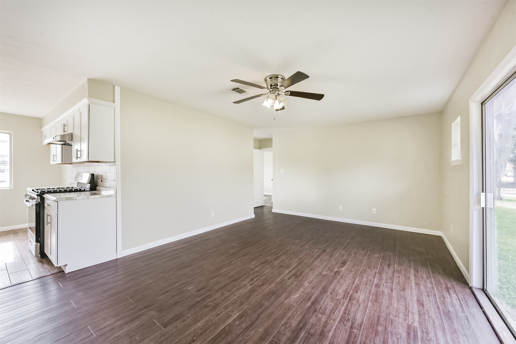 2390 Sarah Street Beaumont, TX 77705 - Photo 5 of 42 This photo showcases a bright, open-concept living area with wood-style flooring. The space includes a modern kitchen with white cabinetry and stainless steel appliances, a ceiling fan, and large sliding glass doors that lead to an outdoor area.