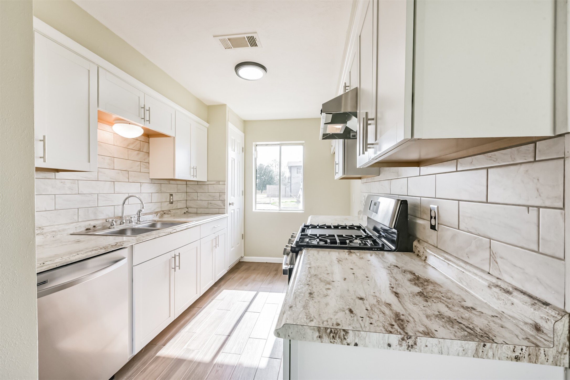 2390 Sarah Street Beaumont, TX 77705 - Photo 10 of 42 Bright, modern kitchen with white cabinets, stainless steel appliances, and a gas stove. The space features a tiled backsplash, marble-patterned countertops, and ample natural light from a window at the end.
