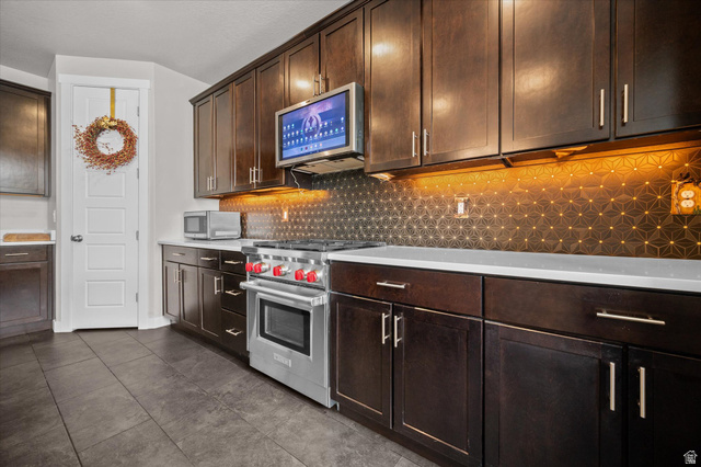 3359 Eurasian Crane Road Clinton, UT 84015 - Photo 8 of 71 Kitchen featuring dark wood finish cabinetry, stainless steel appliances, decorative backsplash, dark tile patterned floors, and light stone counters
