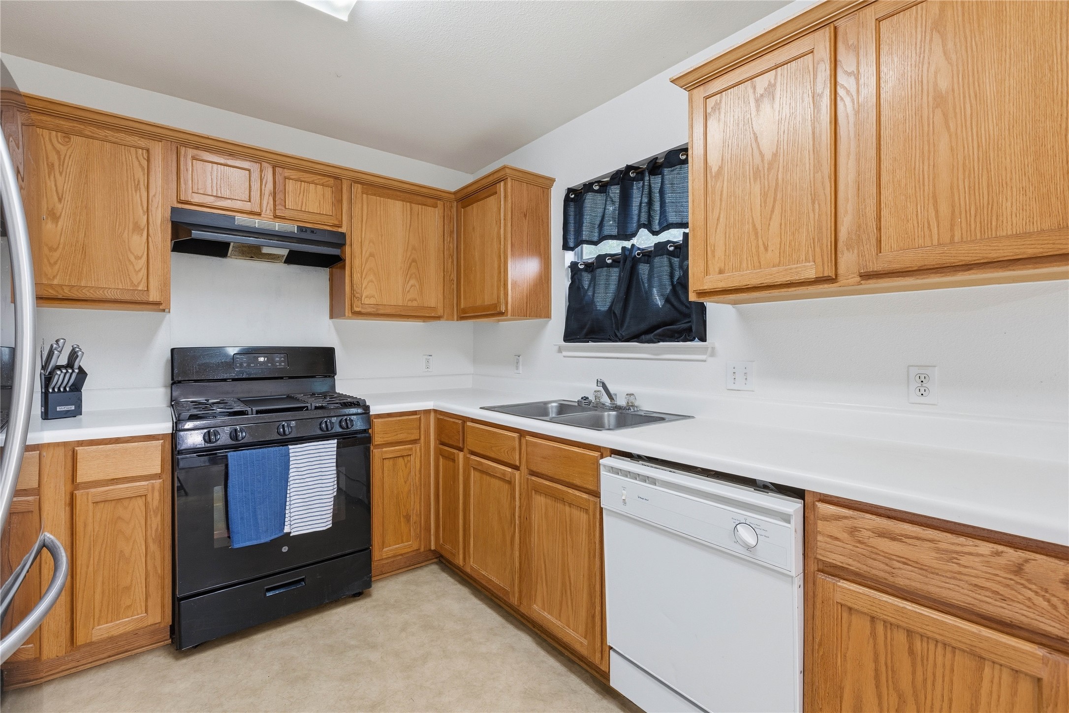 16112 Hamilton Point Circle Manor, TX 78653 - Photo 19 of 27 Kitchen featuring wood cabinetry, white countertops, a black range with an overhead ventilation hood, a double-basin stainless steel sink, and a white dishwasher