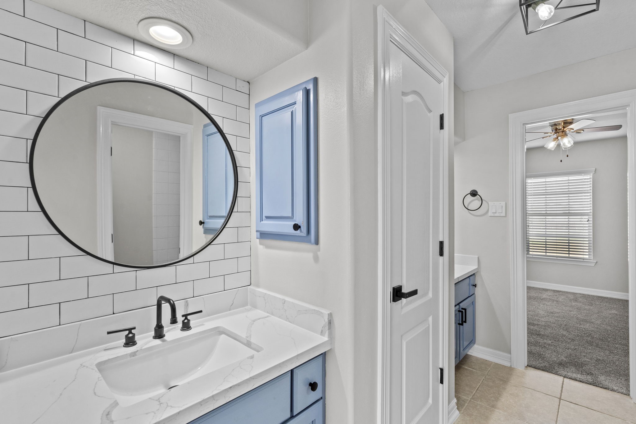 2730 Lawrence Road Kemah, TX 77565 - Photo 38 of 46 This bathroom features a modern design with a white subway tile backsplash, a round mirror, and a sleek black faucet. Light blue cabinetry adds a pop of color, while an adjacent hallway leads to a carpeted room with a ceiling fan.
