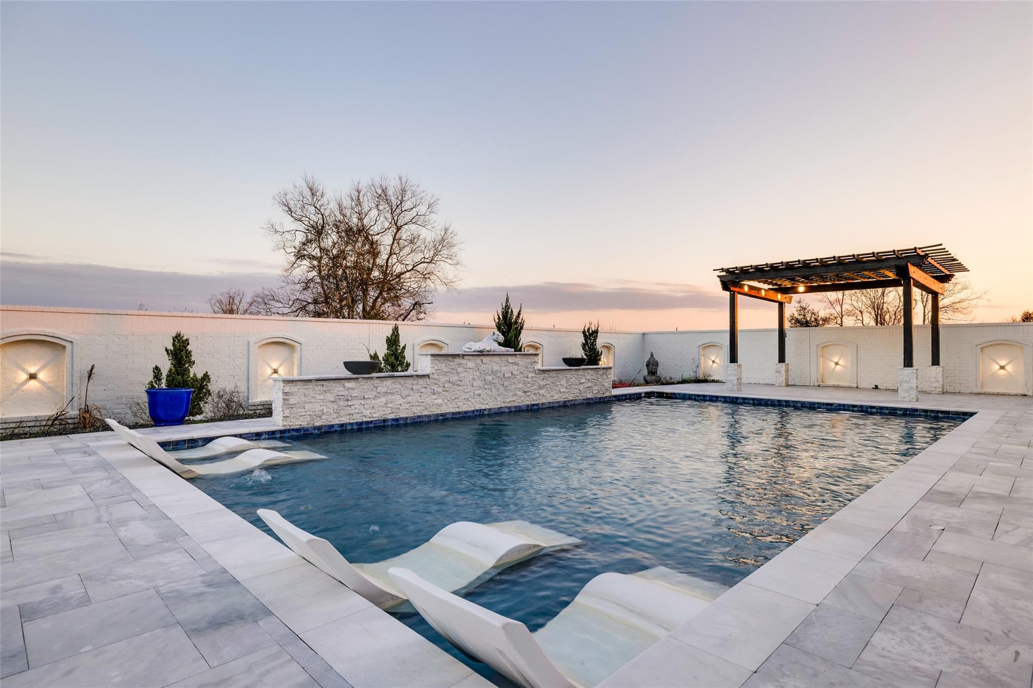4821 Walters Road Richmond, TX 77469 - Photo 6 of 50 This stunning heated luxury pool creates a true resort-style experience with sleek modern design, expansive lounging areas, and breathtaking sunset views. Featuring elegant marble-style decking, in-water chaise loungers, ambient accent lighting, and a striking stone water feature, the space was thoughtfully designed for both relaxation and entertaining. A covered pergola lounge adds the perfect setting for evening gatherings, while the oversized pool offers a seamless blend of comfort & privacy.