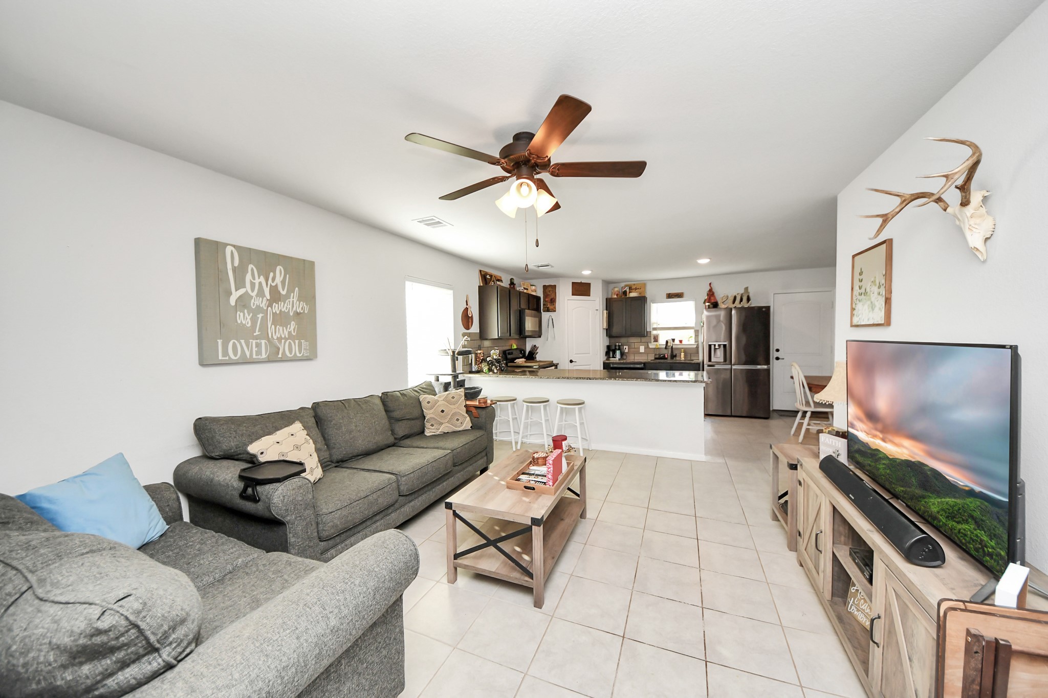 15291 Arrowhead Loop West Willis, TX 77378 - Photo 8 of 28 This open-concept living area features a cozy seating arrangement with neutral-toned sofas and a wooden coffee table. The space flows seamlessly into a modern kitchen equipped with stainless steel appliances and a breakfast bar with stools. The room is well-lit, with a ceiling fan and decor that adds a personal touch. Perfect for comfortable living and entertaining.