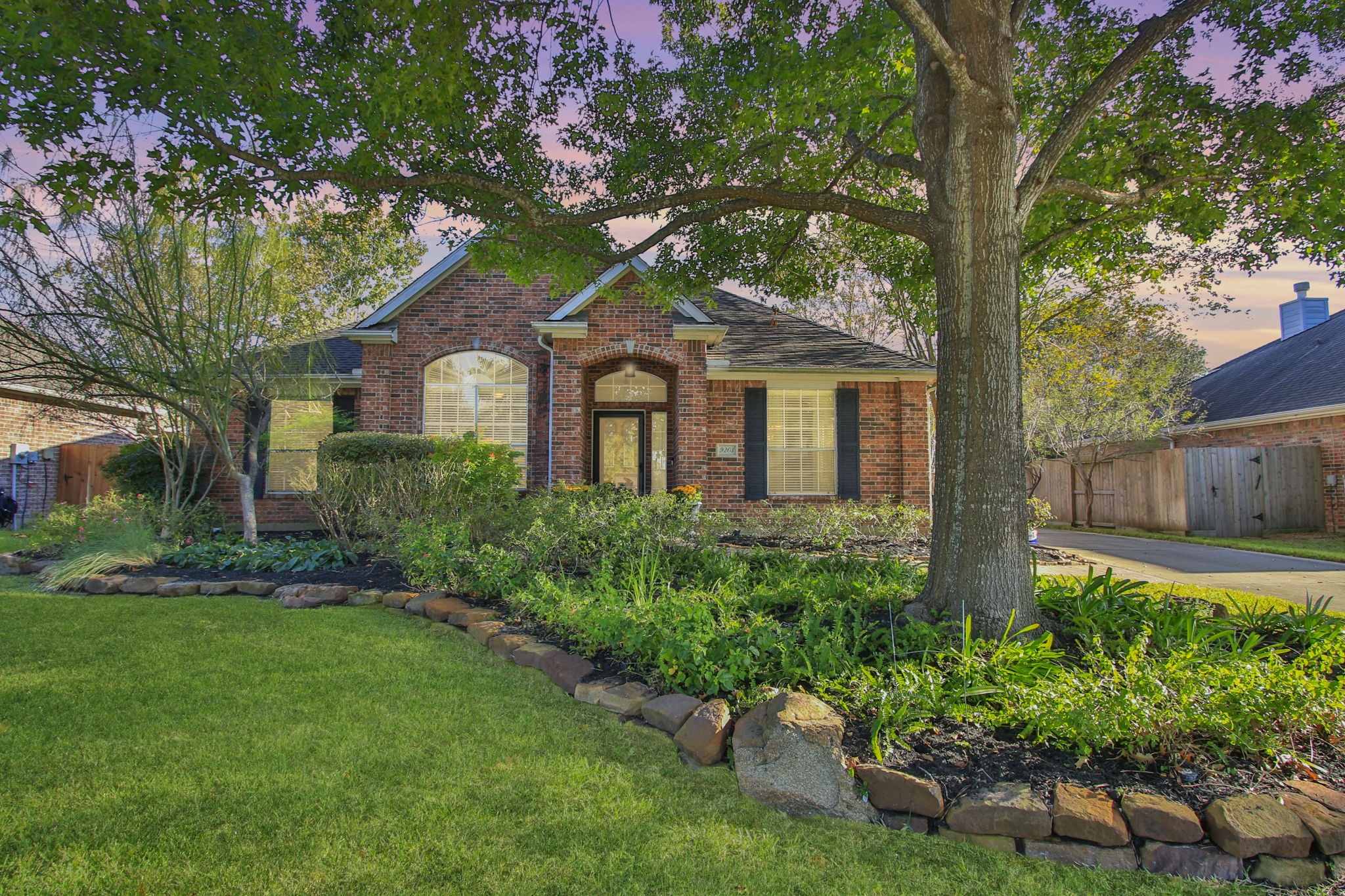 Inviting front elevation framed by mature shade trees and lush landscaping! This charming red brick one-story features classic curb appeal with arched windows, black shutters, and a welcoming covered entry.