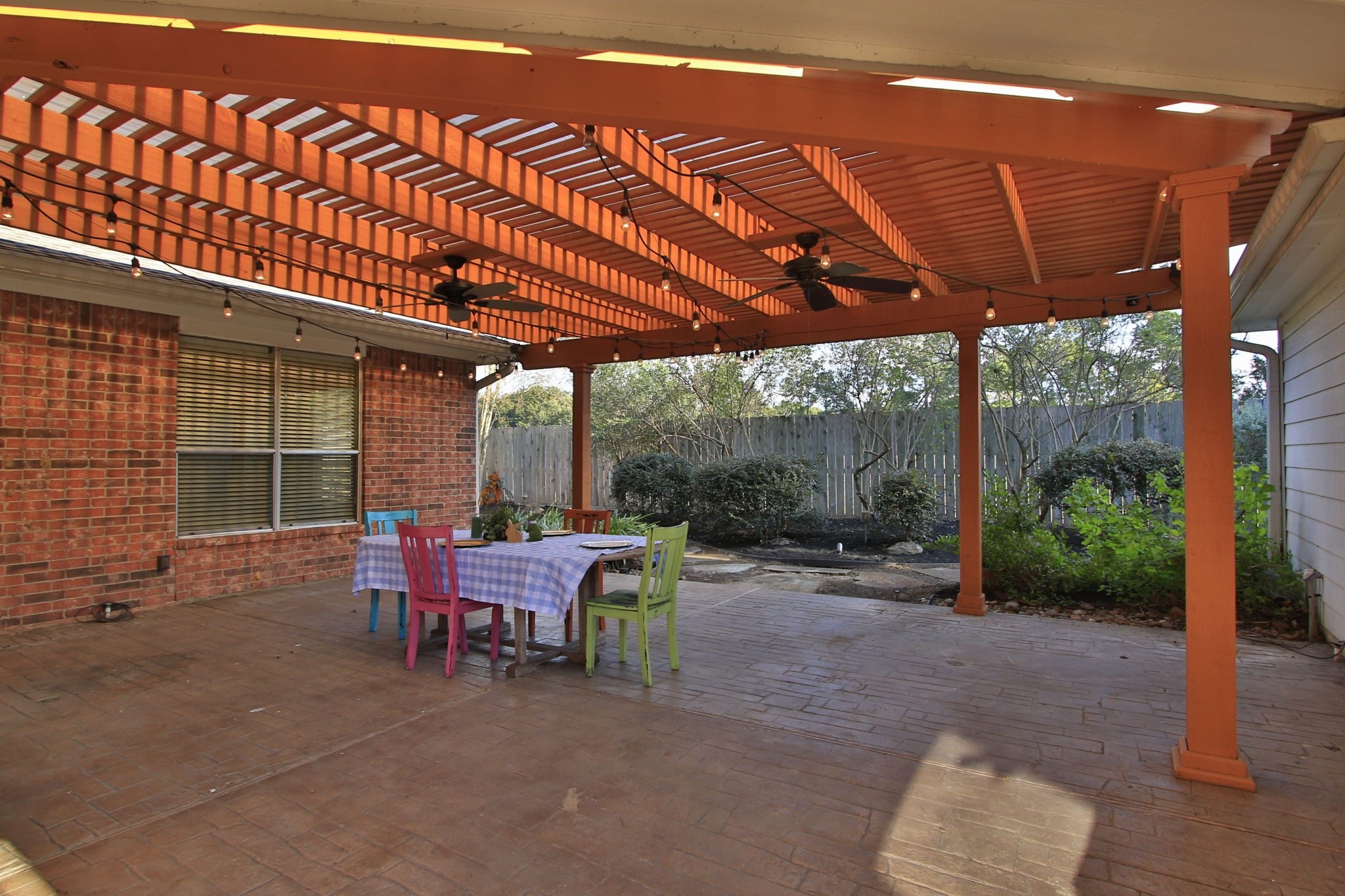 9203 Memorial Pines Way Spring, TX 77379 - Photo 36 of 45 Charming outdoor patio featuring a wooden pergola with string lights and ceiling fans. The space has a large stamped concrete floor, surrounded by a brick exterior and lush greenery, perfect for relaxation and entertaining.