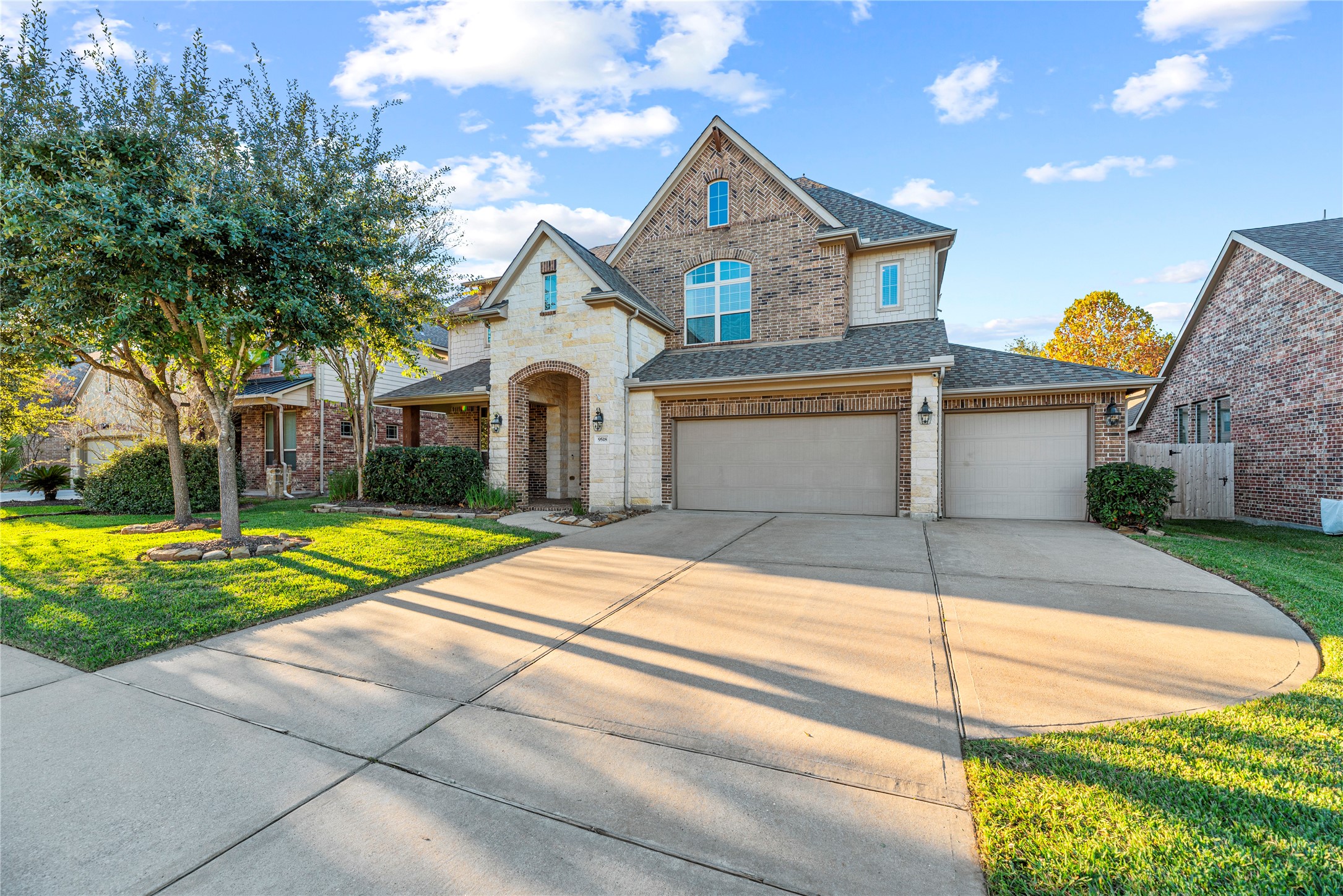 9518 Three Stone Lane Tomball, TX 77375 - Photo 4 of 33 A charming and sophisticated two-story home with a mix of brick and stone exterior. The property features a well-manicured front lawn, mature trees, and landscaped garden beds. The house has a welcoming arched entryway and a three-car across garage, creating an inviting curb appeal.