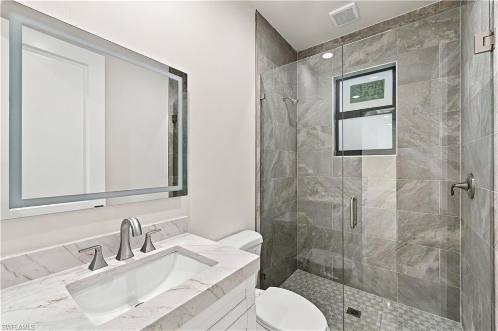 183 5th Street Southwest Naples, FL 34117 - Photo 22 of 40 Backlit vanity mirror, undermount sink, stone countertop, glass-enclosed shower, and tiled shower walls