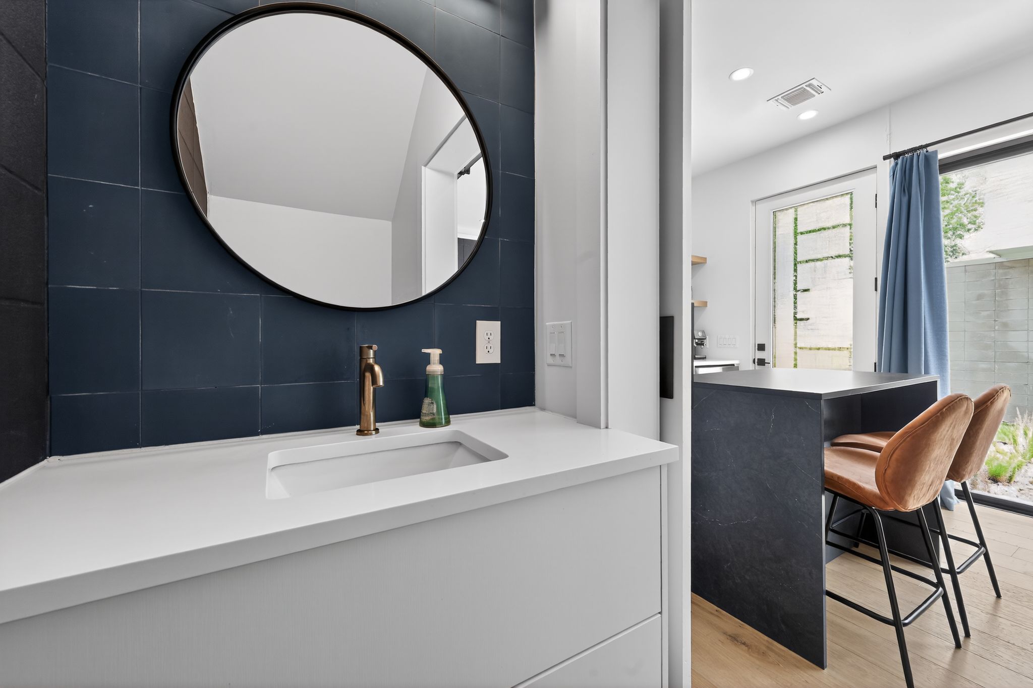 1206 Luna Street, Unit 2 Austin, TX 78721 - Photo 25 of 30 Vanity area featuring a round mirror, a white countertop with an integrated sink, and a gold-toned faucet, set against a dark tiled wall