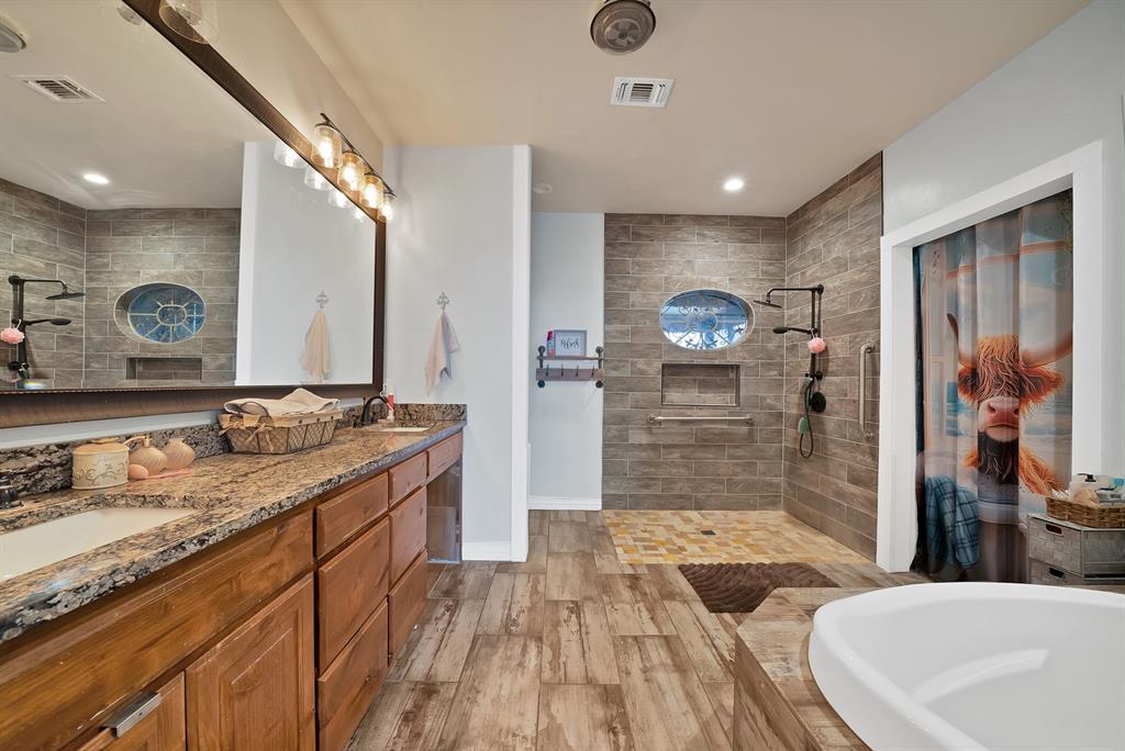100 Shadle Ridge Court Poolville, TX 76487 - Photo 18 of 40 Spacious bathroom featuring a dual vanity with granite-style countertops, wood-finish flooring, a tiled walk-in shower with an oval window, and a built-in tub