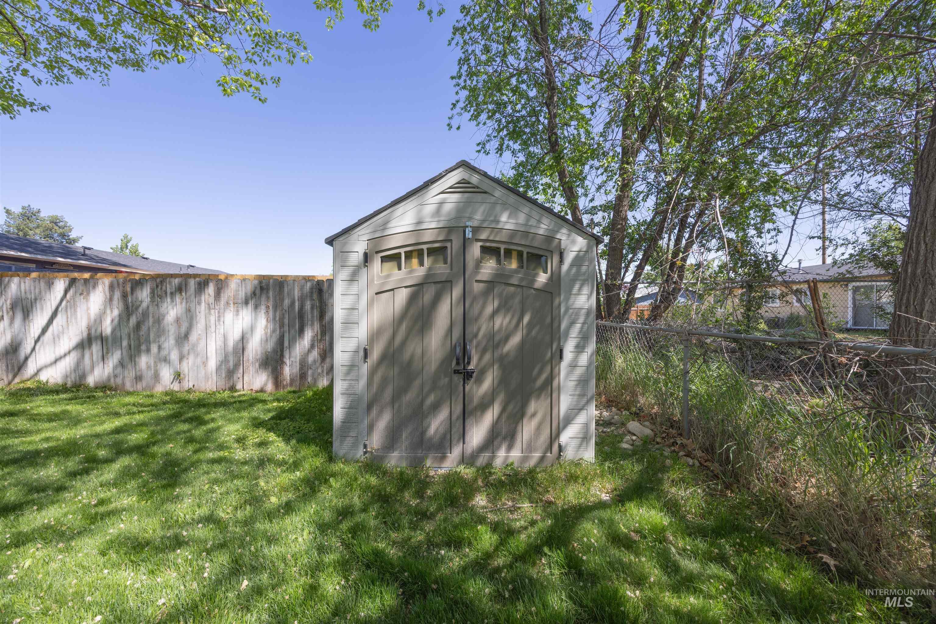 9 Villa Drive Middleton, ID 83644 - Photo 25 of 33 Backyard storage shed with a light gray facade, double doors, and clear panel accents, situated on a grassy lawn with mature trees and a wood fence border