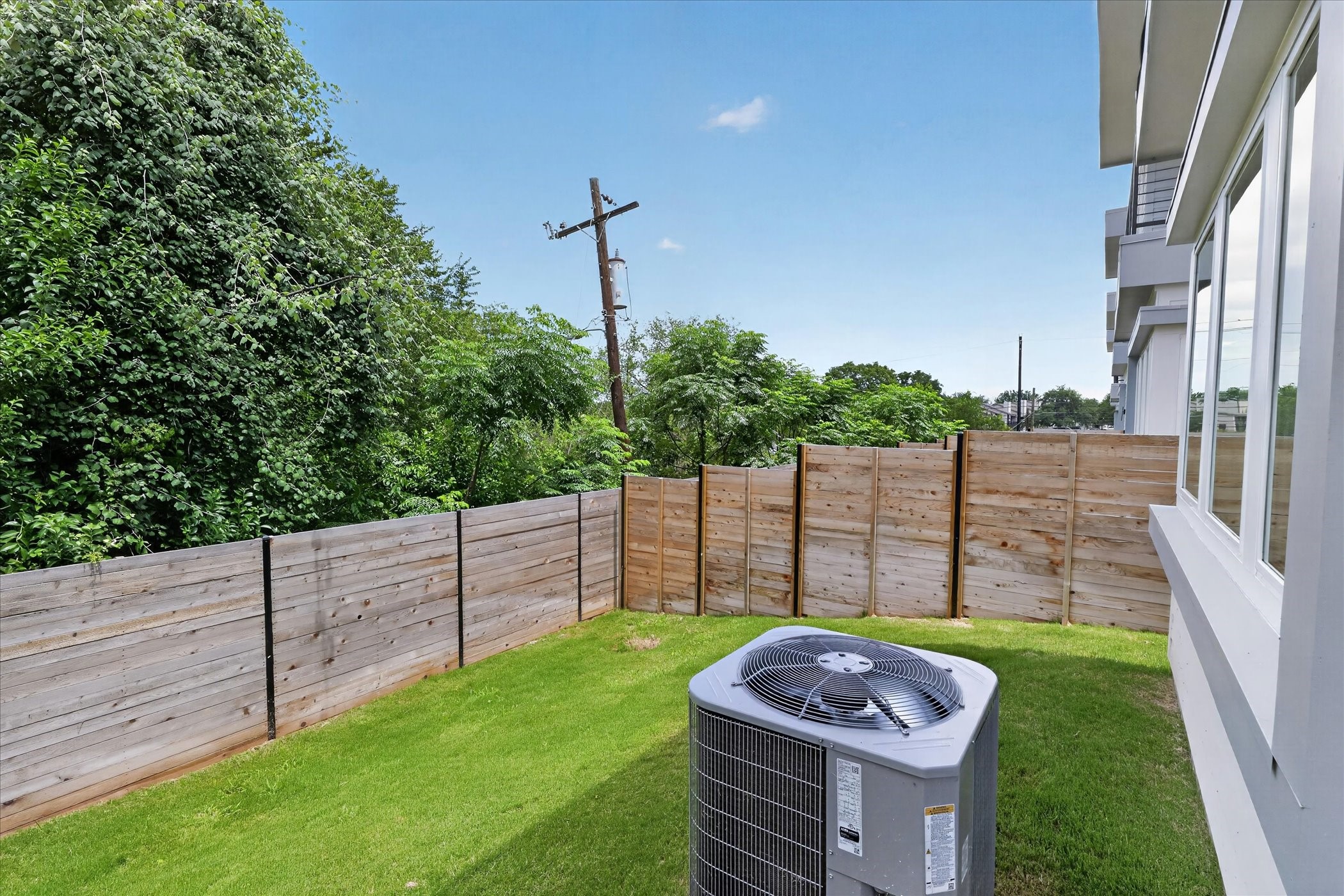 2000 Riverside Farms Road, Unit 8101 Austin, TX 78741 - Photo 38 of 41 This photo shows a small backyard with lush green grass, bordered by a wooden fence. The area features an outdoor HVAC unit and is adjacent to a modern building with large windows. Trees and greenery are visible beyond the fence, providing a natural backdrop.