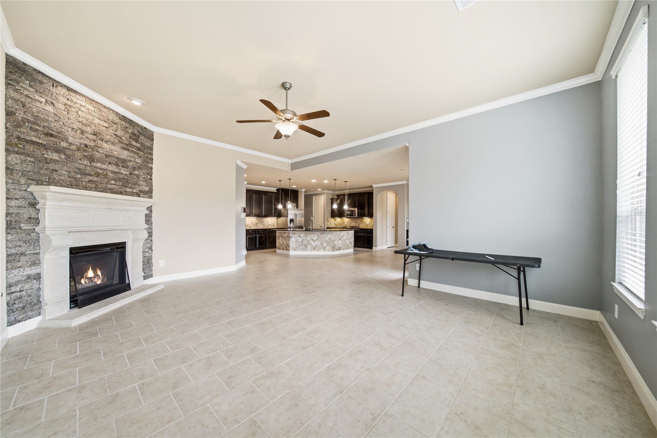 5303 Mesquite Willow Lane Rosharon, TX 77583 - Photo 11 of 32 Modern and classic gas fireplace with a stone accent wall, tile flooring, and an open layout leading to a stylish kitchen with dark cabinetry and a large island. Natural light fills the room through large windows, creating a warm and inviting atmosphere.