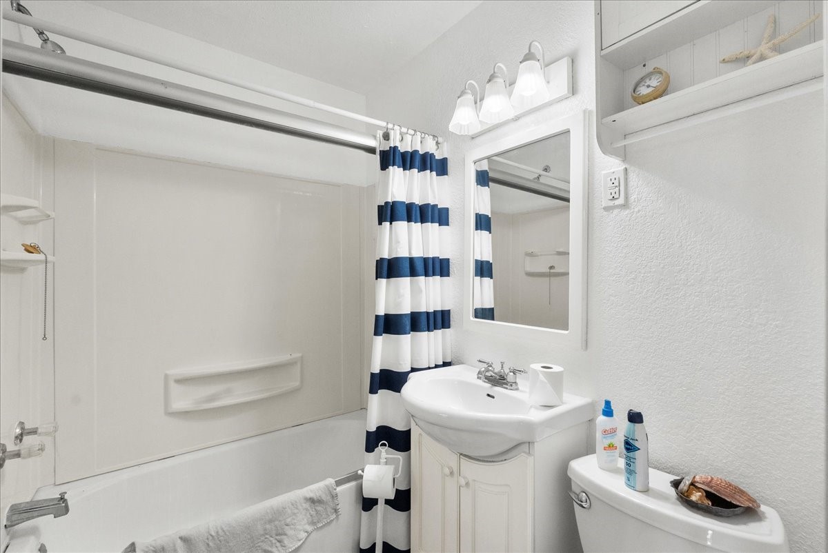 819 Seashell Drive Surfside Beach, TX 77541 - Photo 11 of 27 This bathroom features a clean, bright design with a white and navy striped shower curtain, compact vanity, and overhead lighting. It includes a bathtub with built-in shelving and a small shelf above the toilet for extra storage.