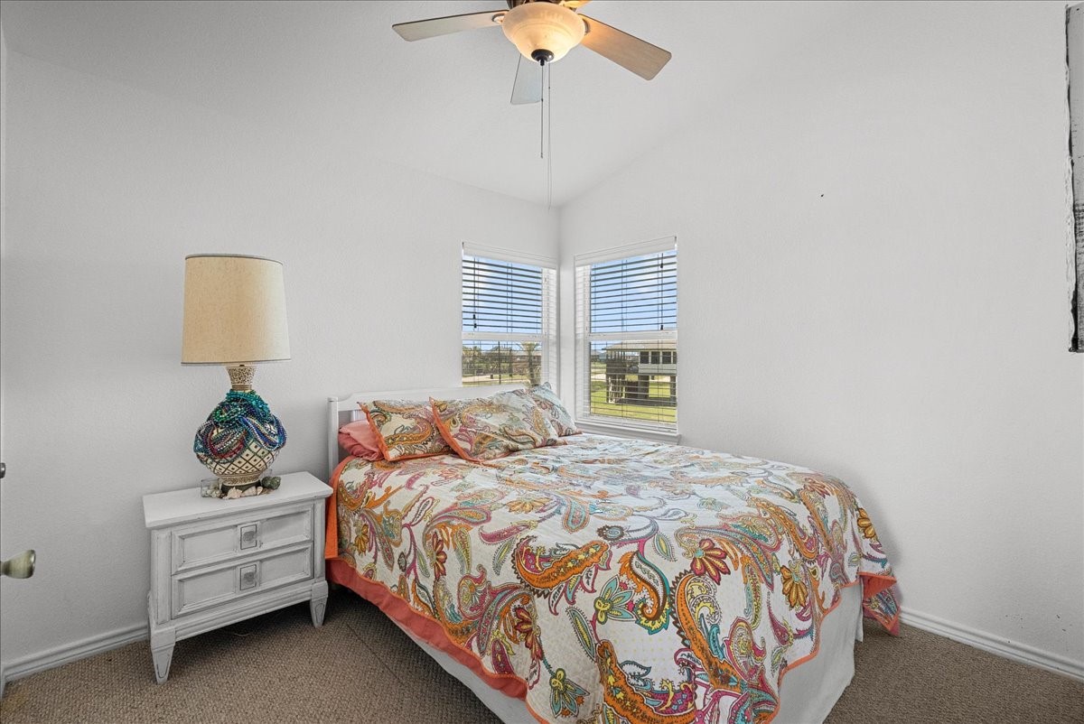 819 Seashell Drive Surfside Beach, TX 77541 - Photo 12 of 27 Cozy bedroom with neutral walls, featuring a colorful quilted bedspread. Natural light streams through two corner windows, while a ceiling fan provides comfort. A unique lamp sits on a white bedside table, adding charm to the space.