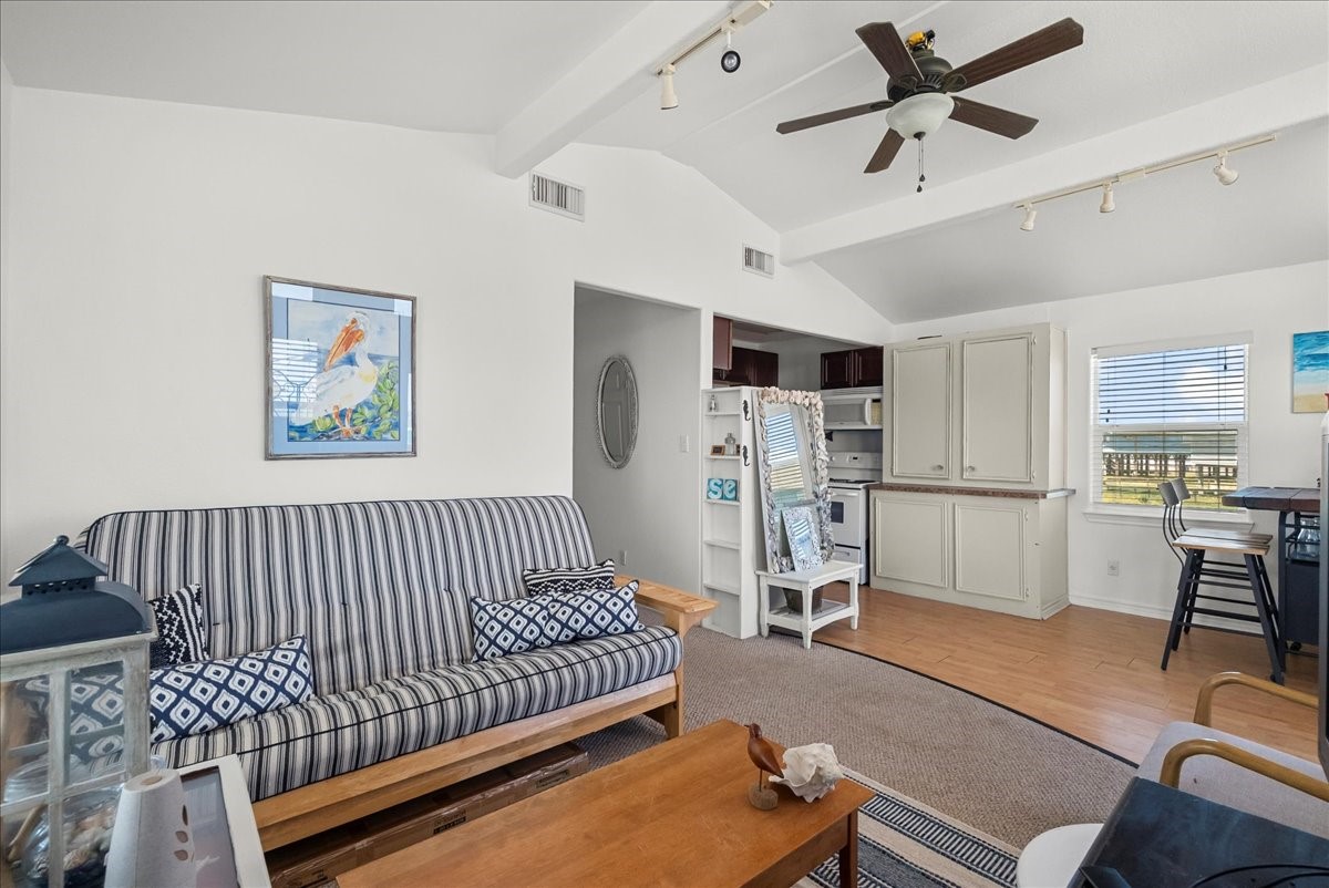 819 Seashell Drive Surfside Beach, TX 77541 - Photo 19 of 27 This cozy living space features a vaulted ceiling with a ceiling fan, a striped sofa, and a wooden coffee table. It has a compact kitchen area with white cabinetry and a small dining nook by the window, offering a bright and airy feel.