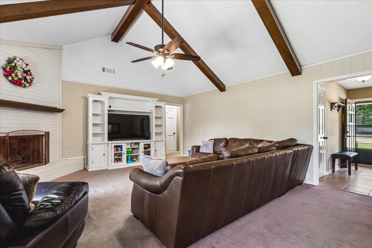 5711 Yale Road Bartlett, TN 38134 - Photo 21 of 29 Vaulted ceiling with exposed wood beams, white painted brick fireplace with wood mantel, built-in media console with shelving, ceiling fan with light fixture, and paneled walls