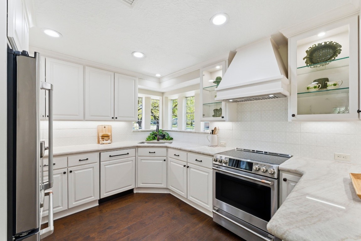 20 Falling Star Road The Woodlands, TX 77381 - Photo 16 of 44 This kitchen features a bright, modern design with white cabinetry, stainless steel appliances, and elegant marble countertops. It includes a sleek range hood, recessed lighting, and dark wood flooring, creating a stylish and functional space.