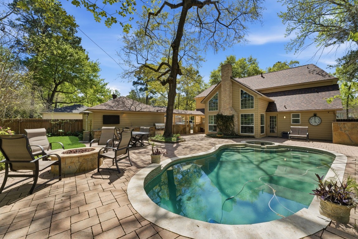 20 Falling Star Road The Woodlands, TX 77381 - Photo 35 of 44 This inviting backyard features a spacious patio with a cozy fire pit, outdoor kitchen, outdoor seating, and a sparkling pool surrounded by lush trees. The charming two-story home has large windows and a stone chimney, offering a perfect blend of relaxation and style.