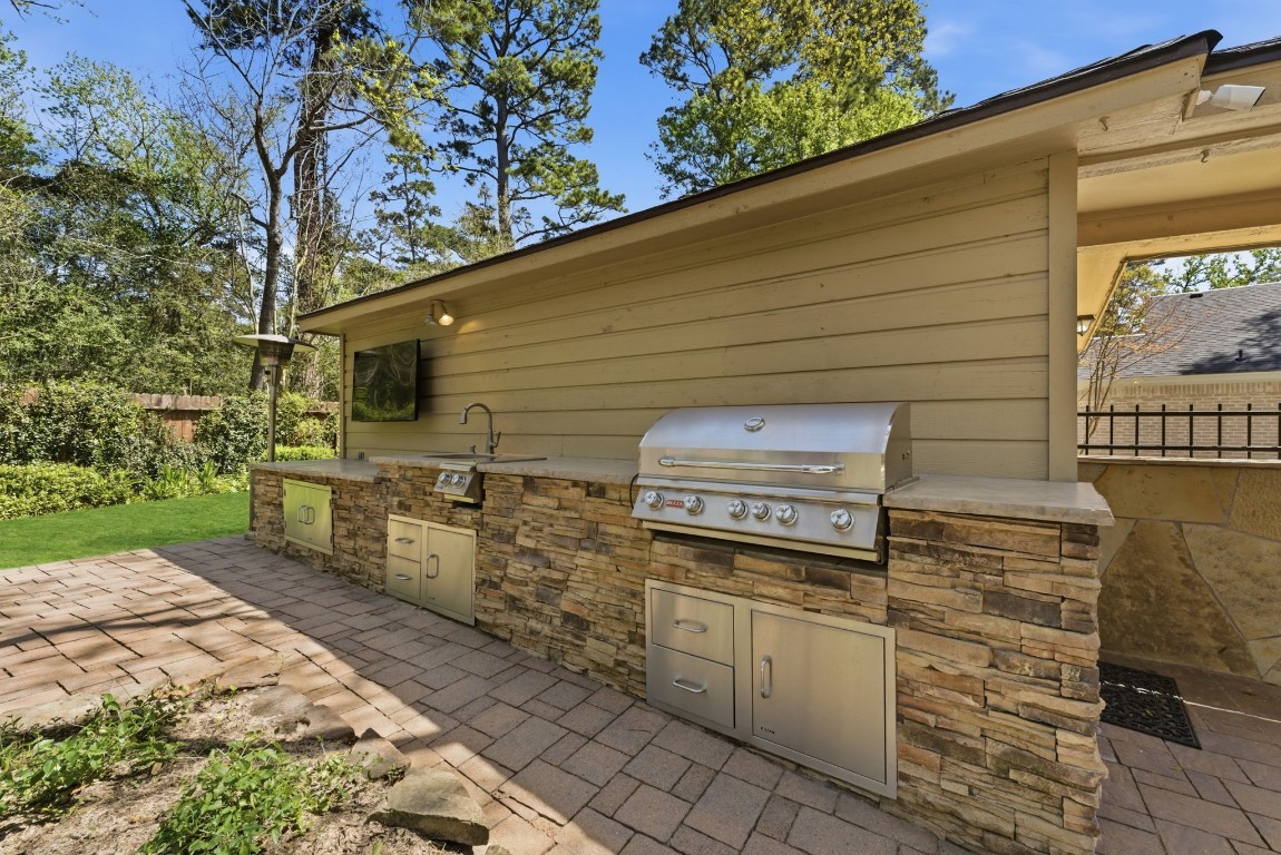 20 Falling Star Road The Woodlands, TX 77381 - Photo 37 of 44 The outdoor kitchen has a built-in stainless steel grill, stone facade, and tiled countertop. It features a sink, storage cabinets, and a mounted TV, set on a spacious patio surrounded by lush greenery, perfect for entertaining.