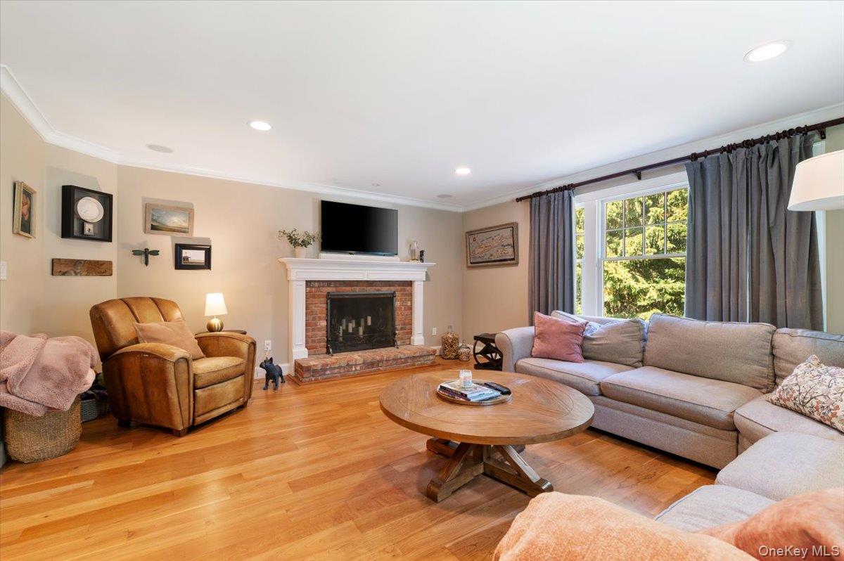 5 Fox Hunt Court Cold Spring Harbor, NY 11724 - Photo 4 of 42 Spacious living area featuring wood-finish flooring, a brick fireplace with a white mantel, recessed lighting, crown molding, and a large window with privacy drapes