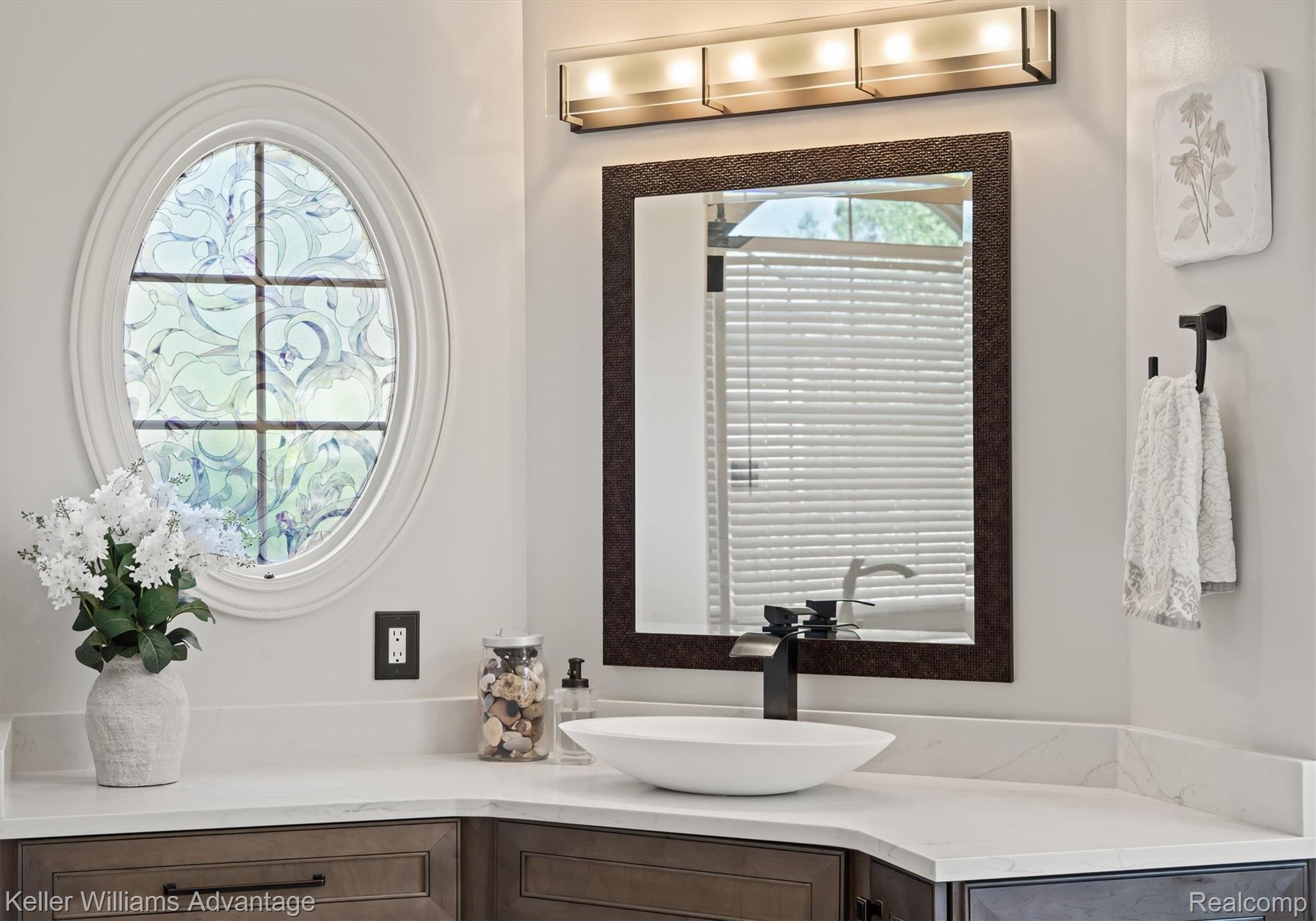 22878 Tree Top Court South Lyon, MI 48178 - Photo 23 of 75 Bathroom vanity featuring a white oval window with decorative glass, a rectangular mirror with an ornate frame, a vessel sink with a matte black faucet, light-toned countertops, and dark wood-finish cabinetry