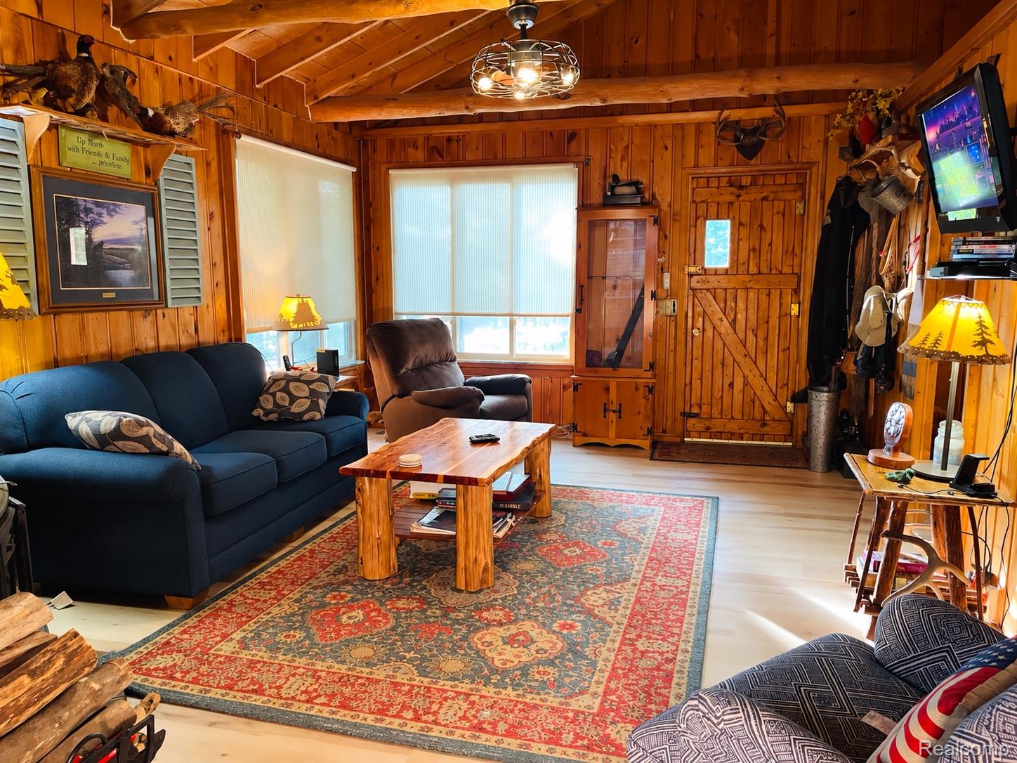 417 Ogemaw Road Rose City, MI 48654 - Photo 5 of 26 Wood-paneled walls and ceiling beams, light wood-finish flooring, multiple windows with roller shades, and a rustic wood plank entry door